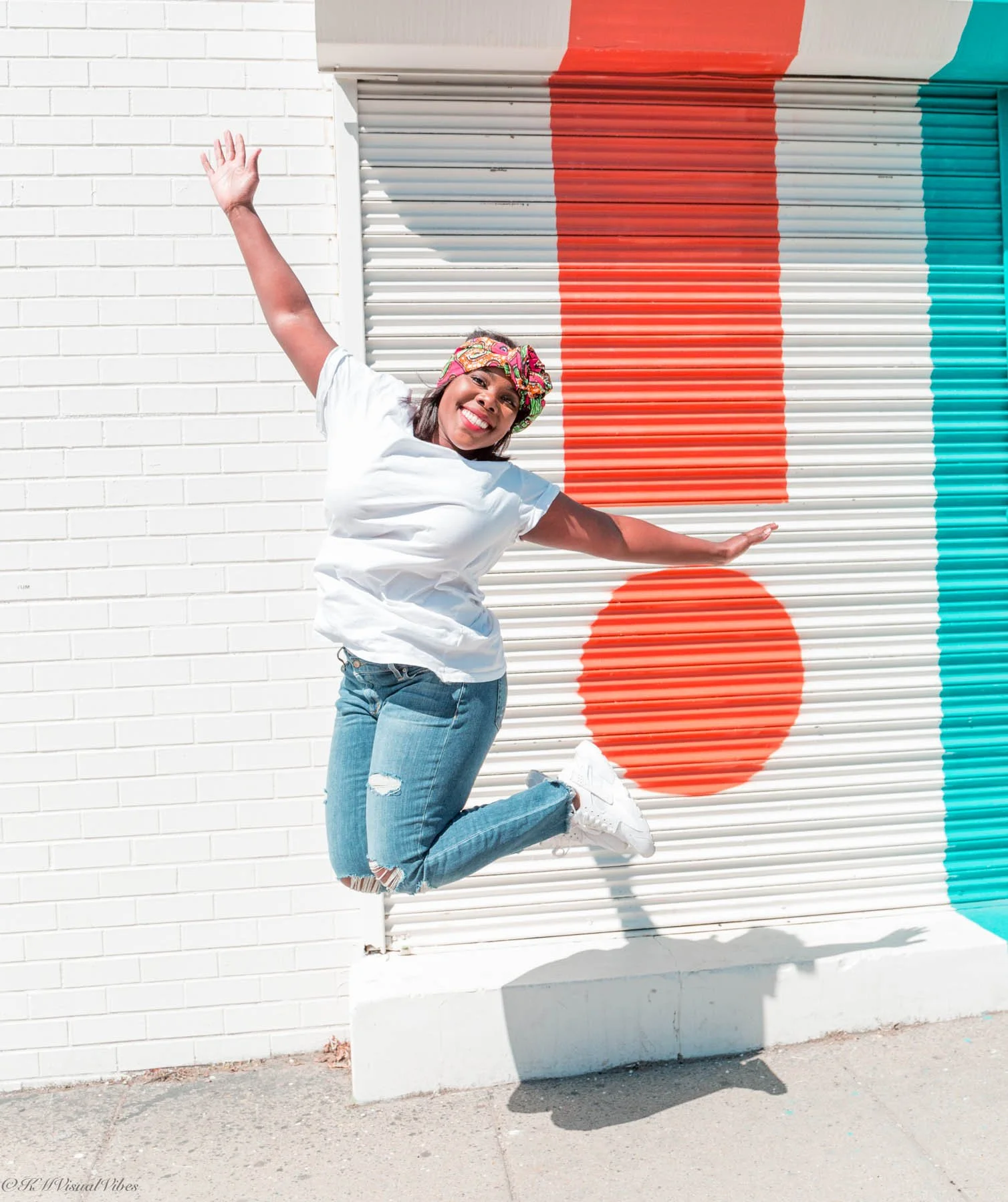 Young woman jumping in front of a colorful mural on a shutter, smiling and wearing casual clothes with a colorful headscarf.