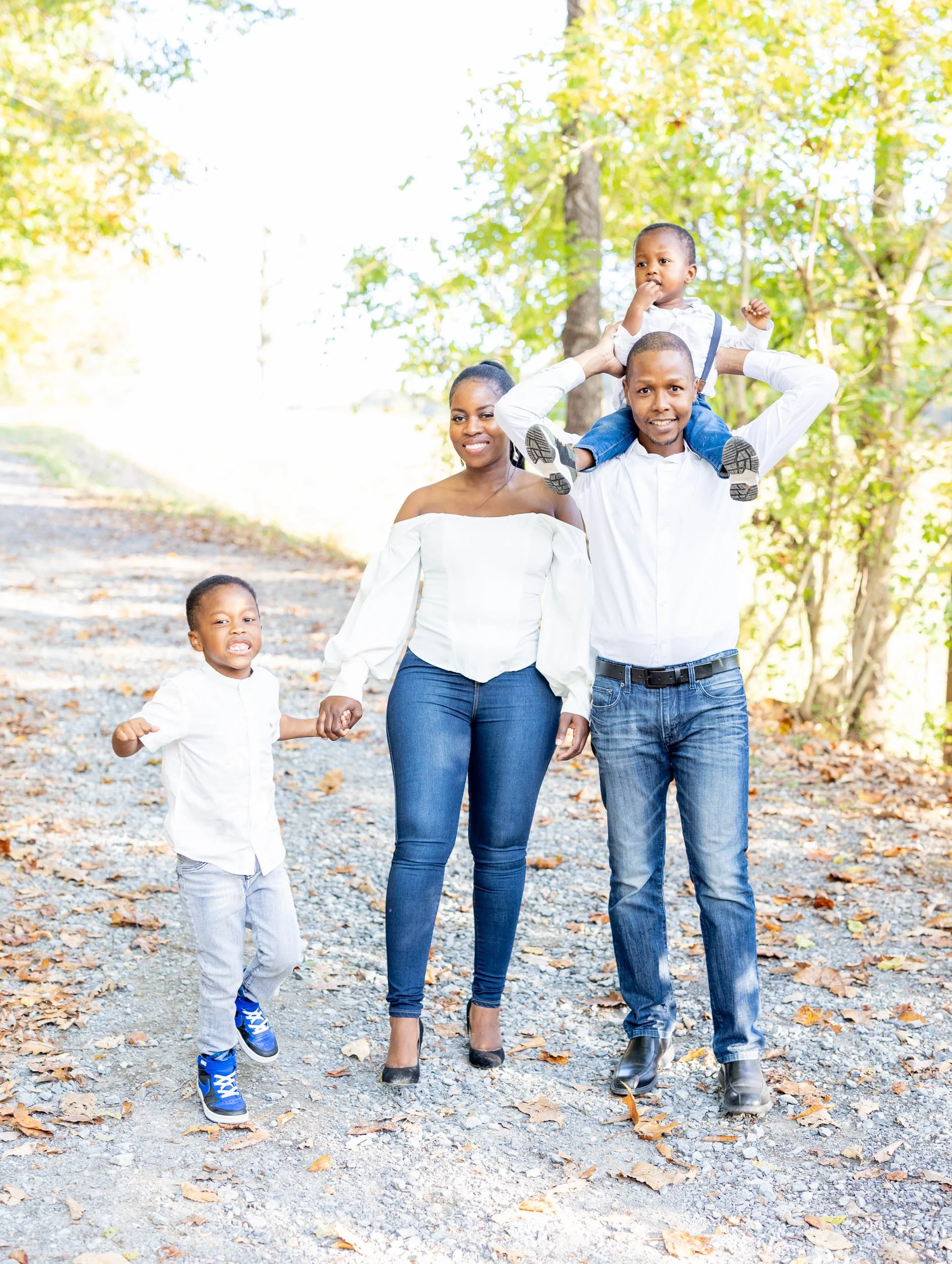 A happy family walking outdoors on a fall day, with green trees and fallen leaves on the ground, a woman, two young boys, and a man carrying a boy on his shoulders.