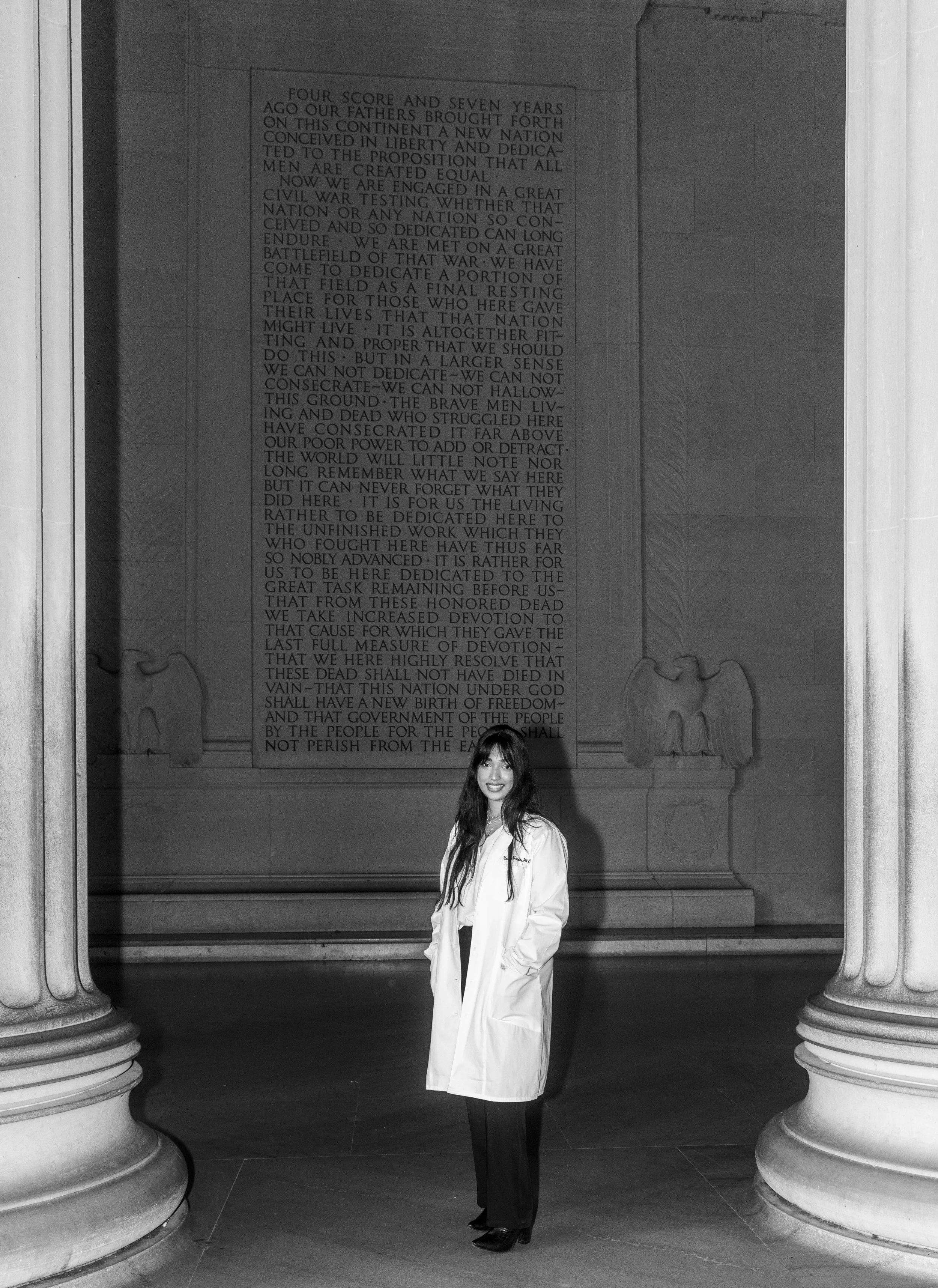 A woman in a white coat standing in front of the Lincoln Memorial in Washington, D.C., with engraved text and large classical columns in the background.