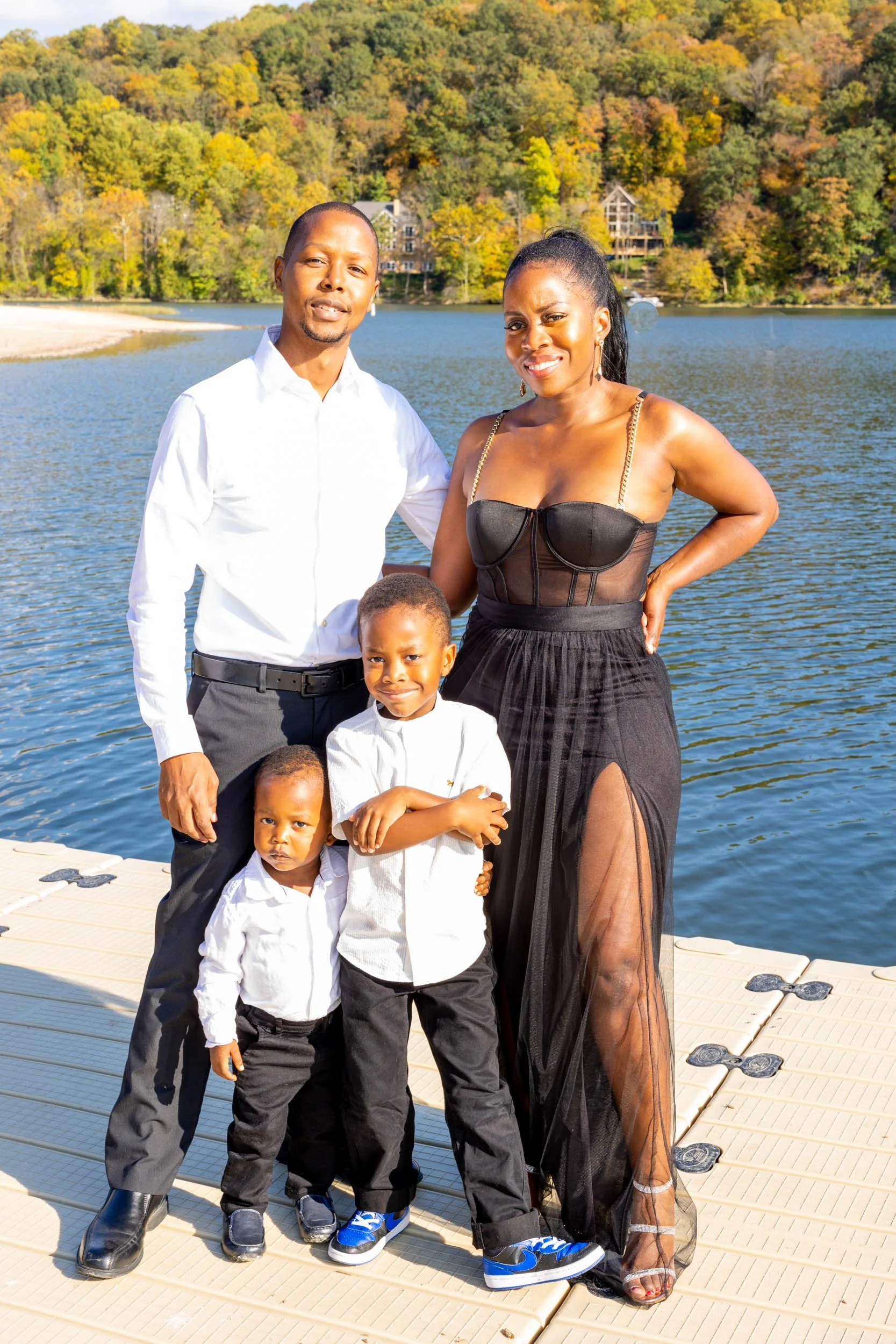 A family of five standing on a dock by a lake surrounded by fall-colored trees. The father is in a white shirt and black pants, the mother in a black dress with sheer panels, and three young boys in white shirts and dark pants.