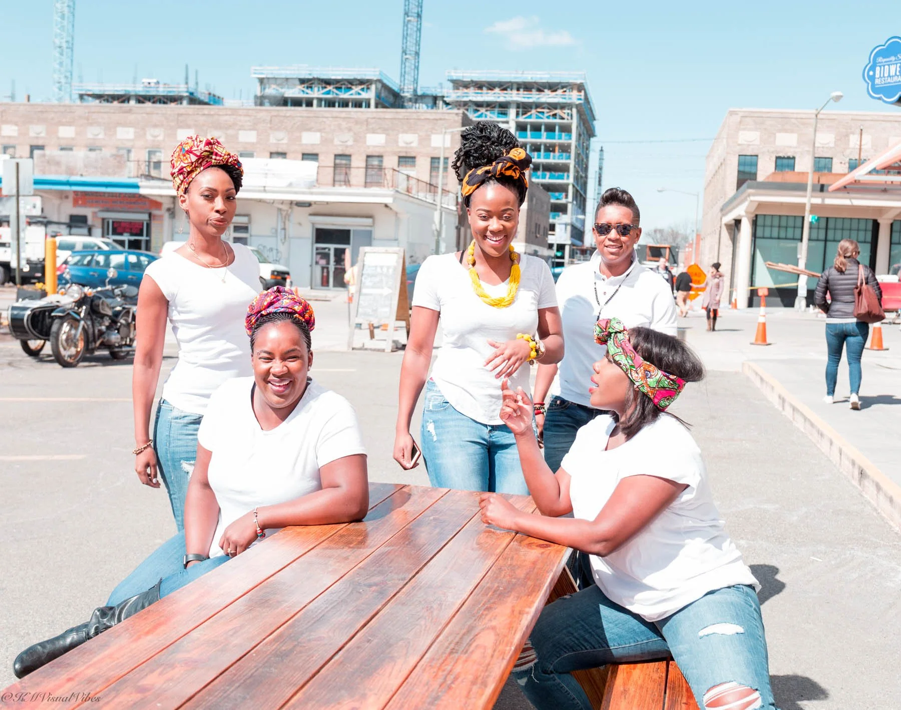 Five women of diverse backgrounds sitting and standing around a wooden table outdoors, talking and smiling on a sunny day, with construction and storefronts in the background.