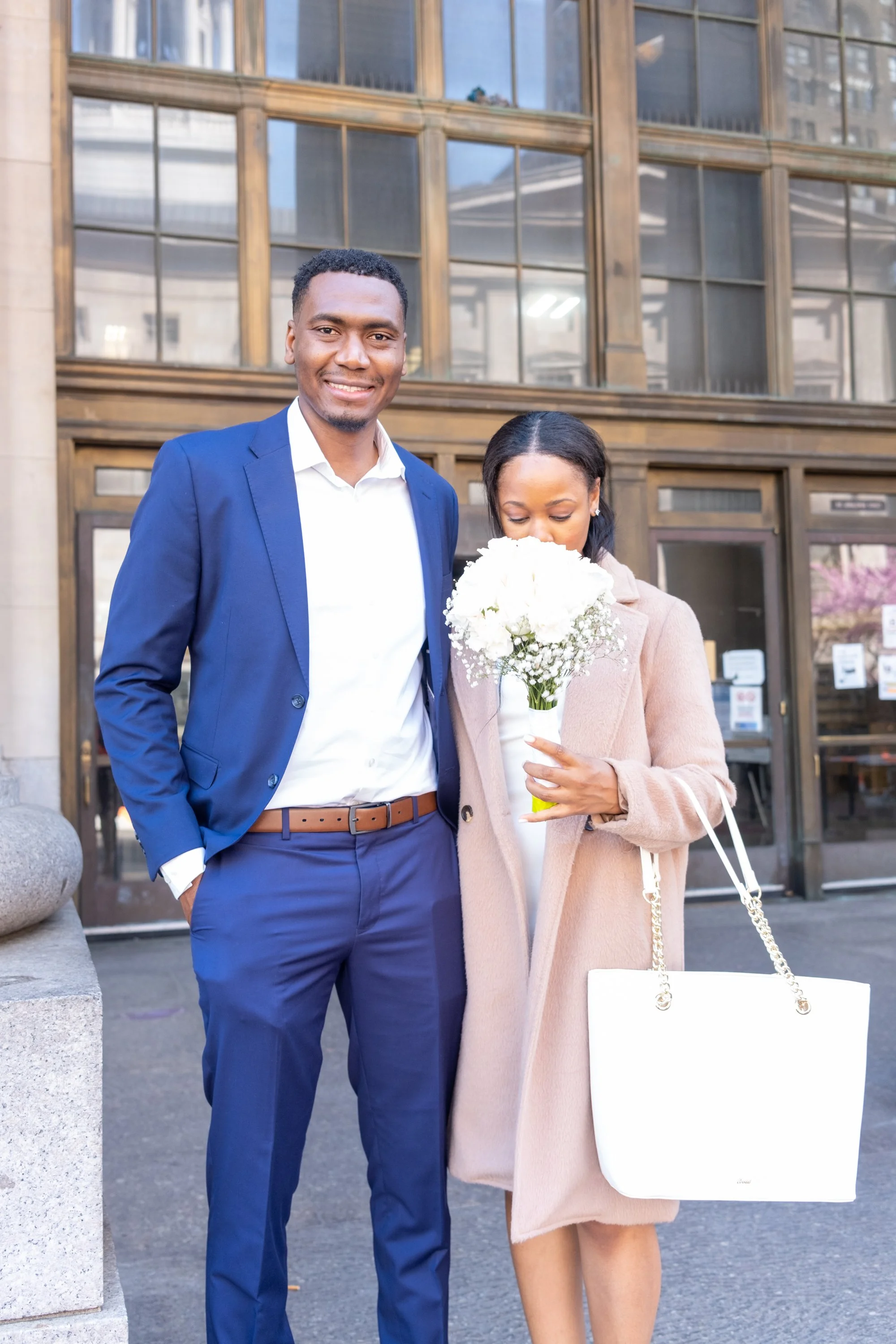 A man in a blue suit and a woman in a beige coat standing outside a building, with the woman holding a bouquet of white flowers.