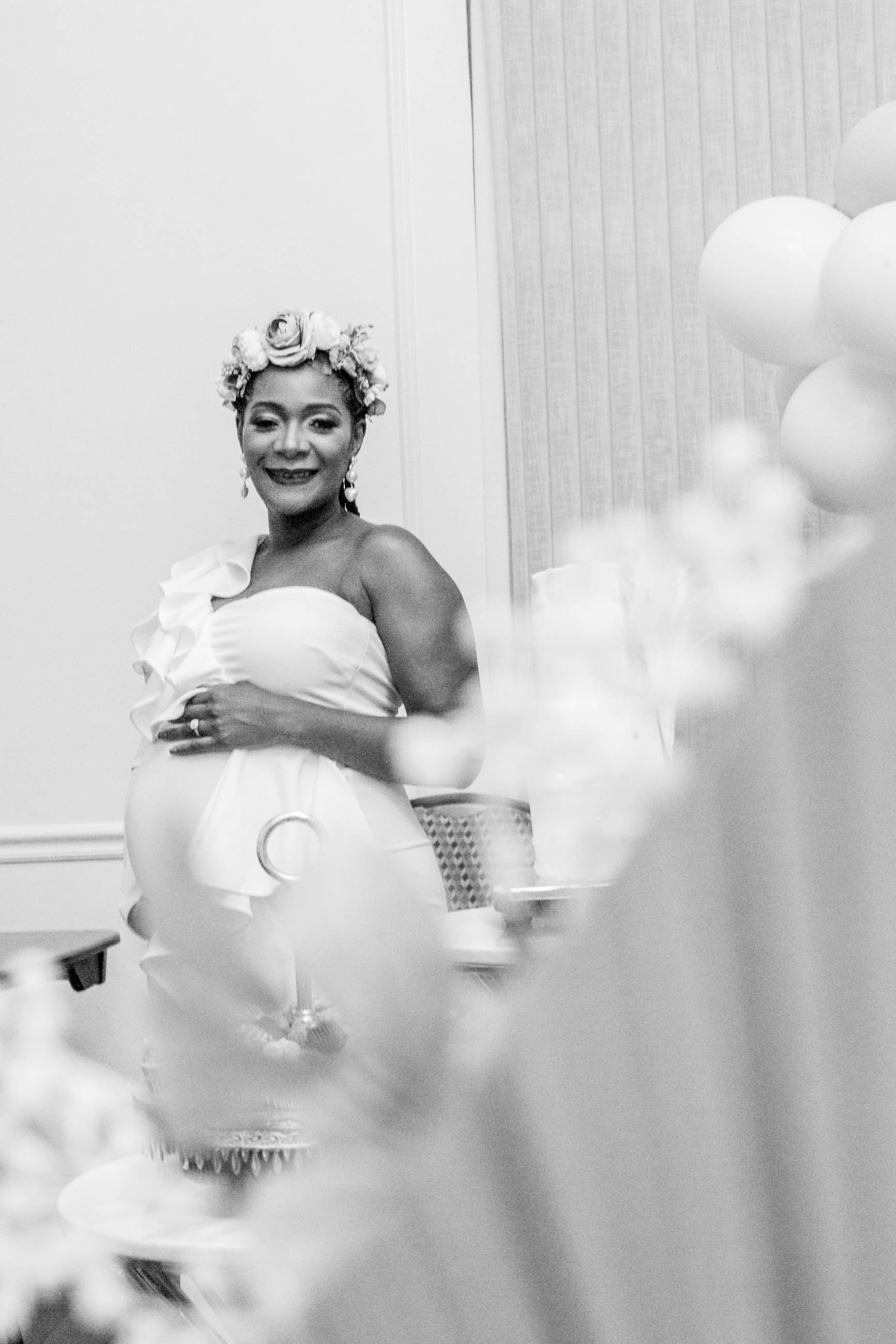 A smiling woman wearing a flower crown and a strapless dress, touching her pregnant belly, at a celebration or party setting.