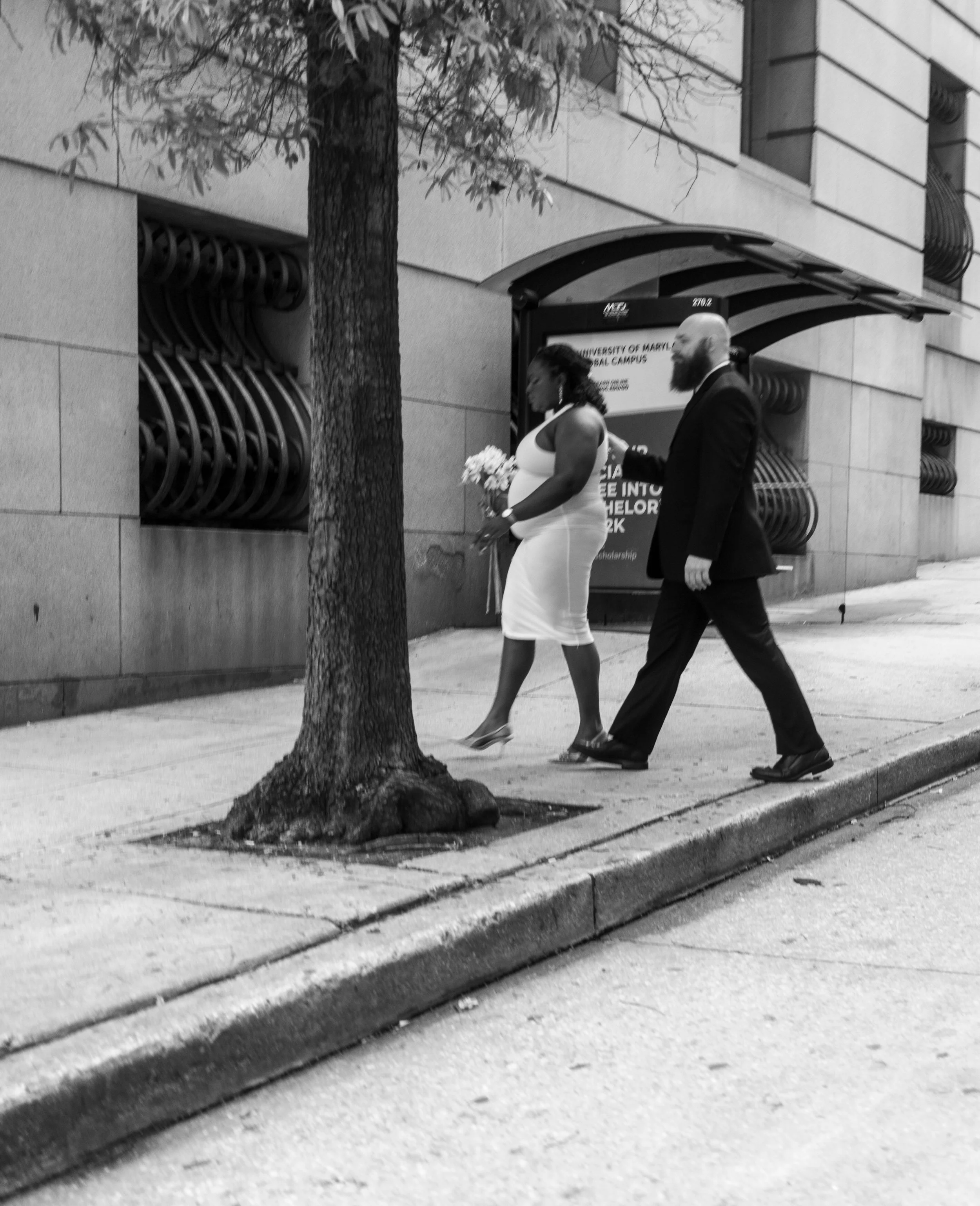 A black-and-white photo of a woman in a white dress holding flowers walking on the sidewalk with a man in a suit walking beside her, near a building with a tree in the foreground.