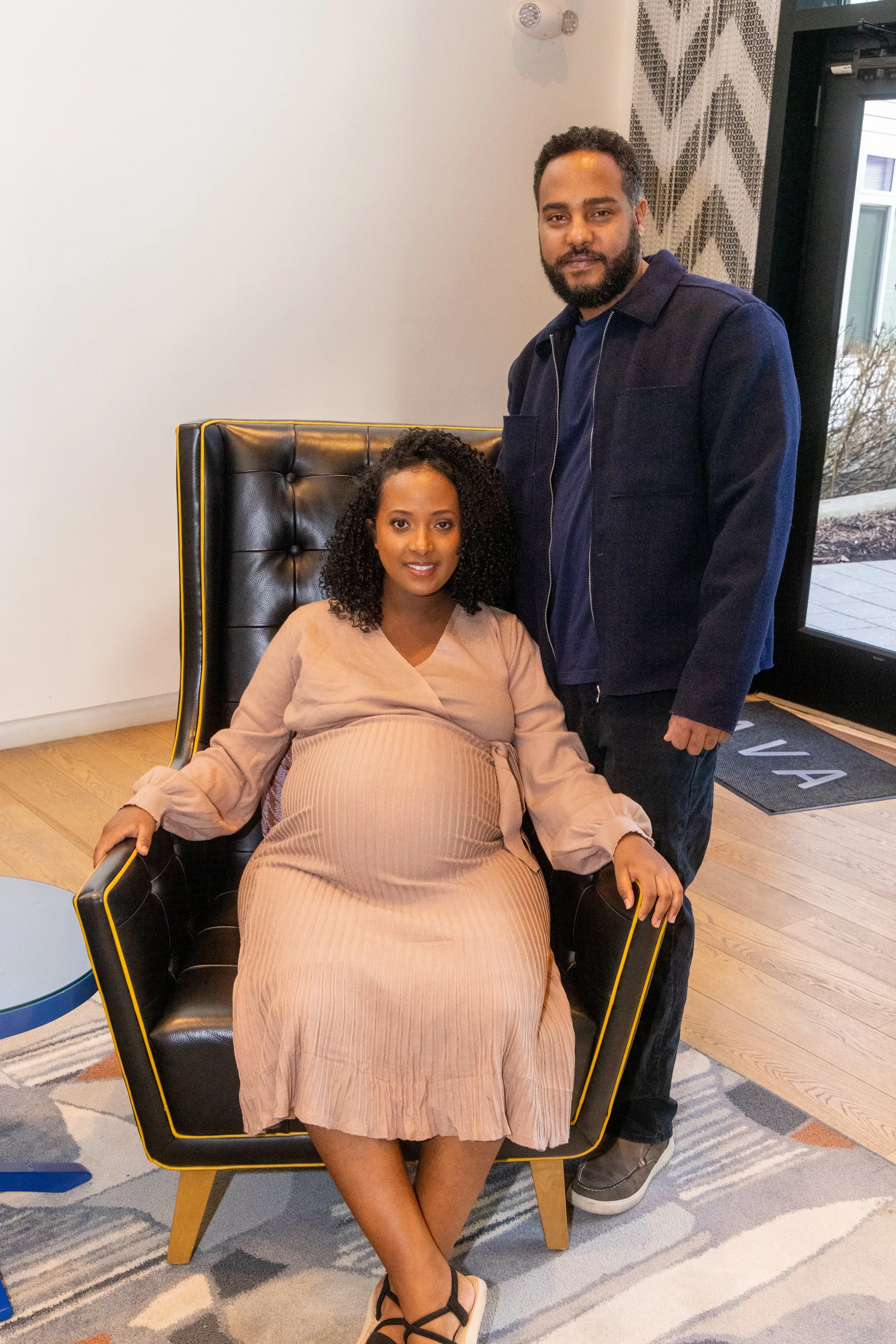A pregnant woman in a beige dress sitting in a black armchair with yellow trim, alongside a standing man, in a modern indoor space with wooden flooring and a large window.