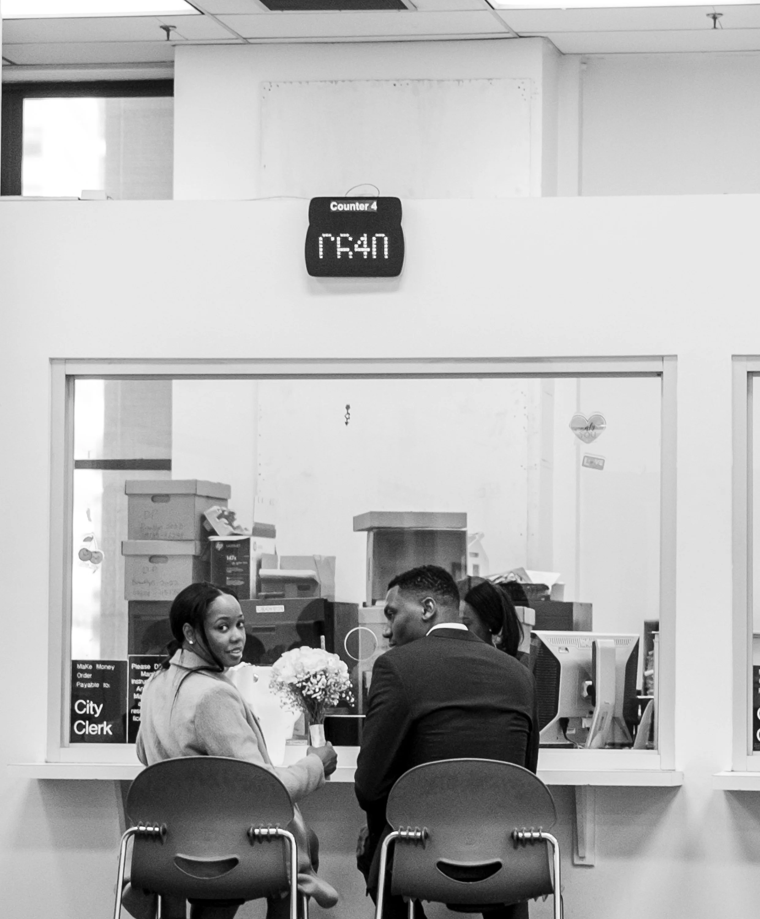 Two people sitting at a counter in an office or government building, with one person holding a bouquet of flowers, seen through a large glass window, with office supplies and computers behind the window.