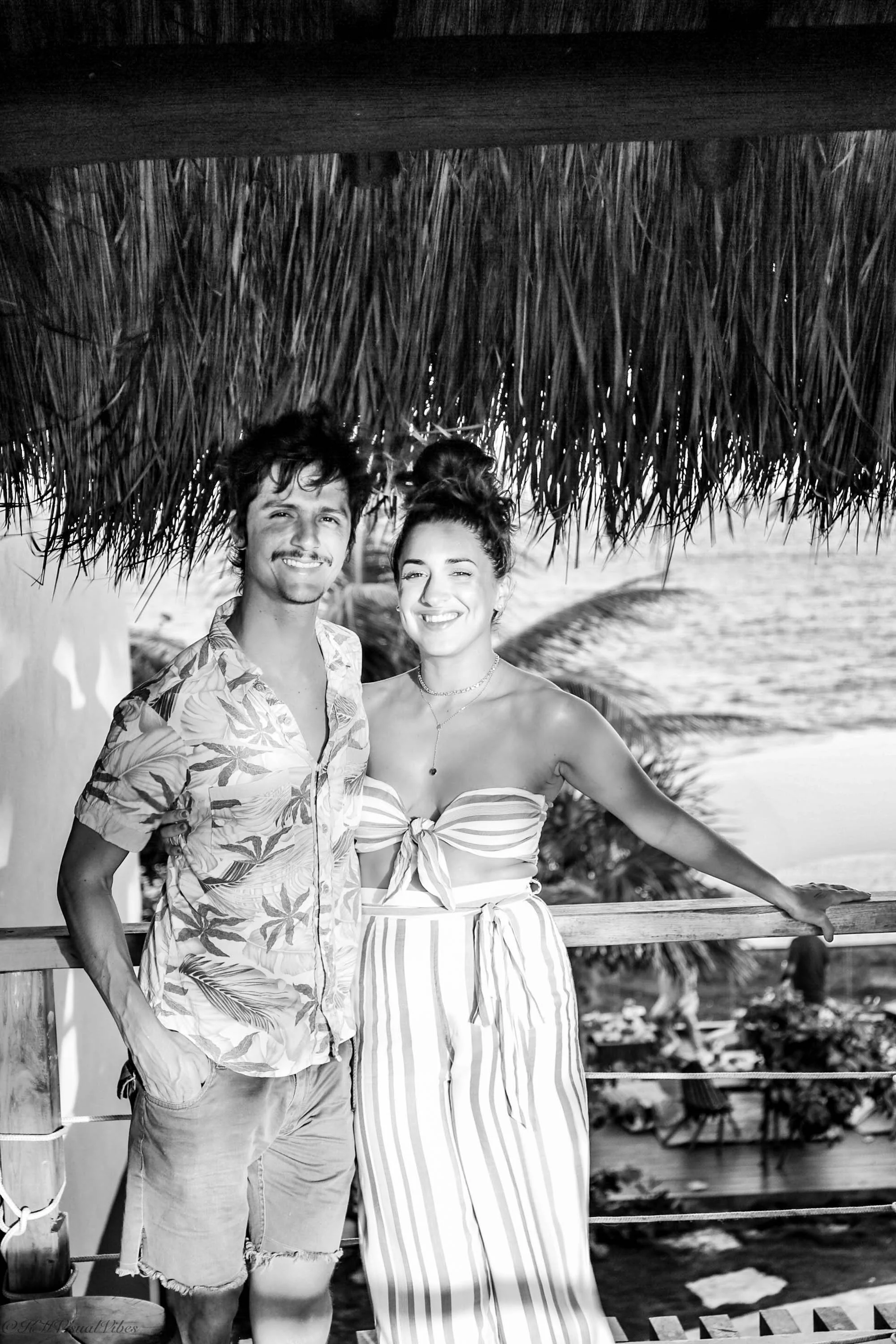 A smiling young man and woman standing under a thatched roof on a balcony with tropical plants and a beach in the background.