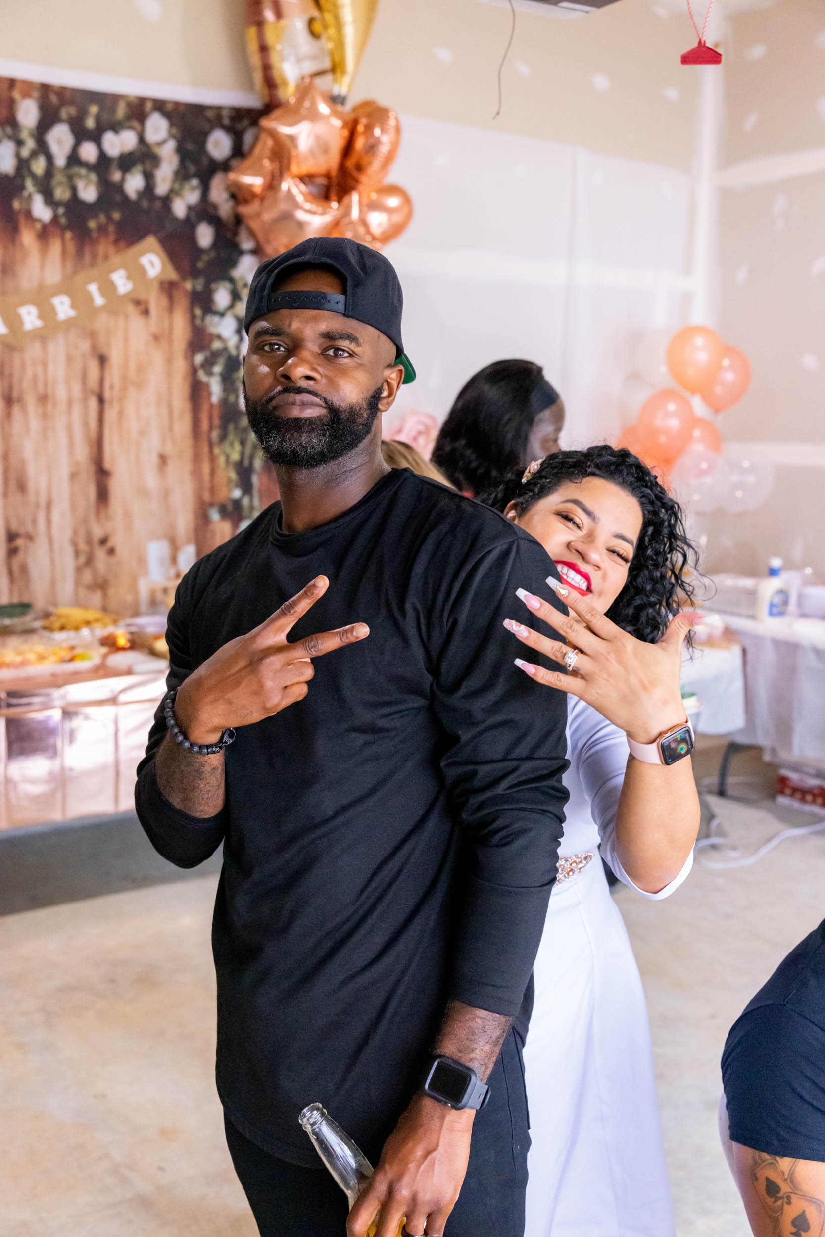 A man in a black shirt and a backwards baseball cap making a peace sign, standing next to a woman with curly hair celebrating with a smile at a party with balloons and background decorations.