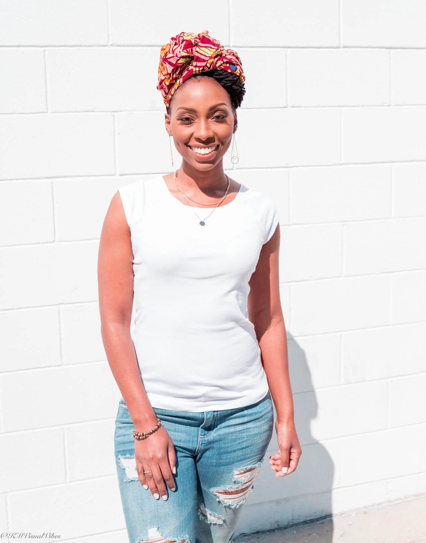 A smiling woman with dark skin, wearing a colorful headwrap, gold jewelry, a white sleeveless top, and ripped jeans, standing in front of a white brick wall.