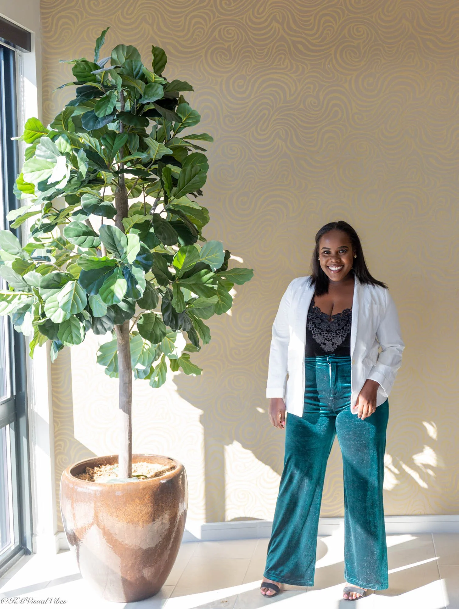 A woman standing next to a large potted indoor plant in a well-lit room with a decorative yellow wall background.
