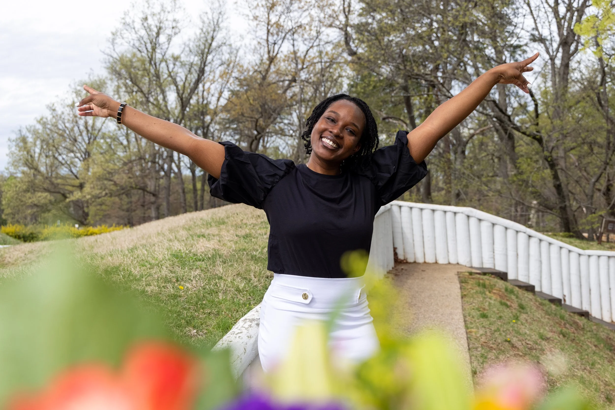 A smiling woman with her arms raised in a park with trees and a curved white railing in the background.