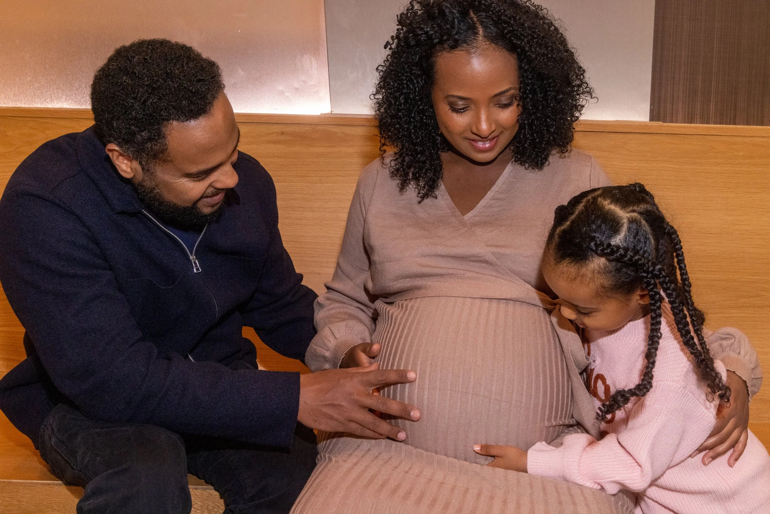 A family of three, including a pregnant woman and her daughter, sitting on a bench, with the daughter and husband touching her pregnant belly and the daughter laughing.