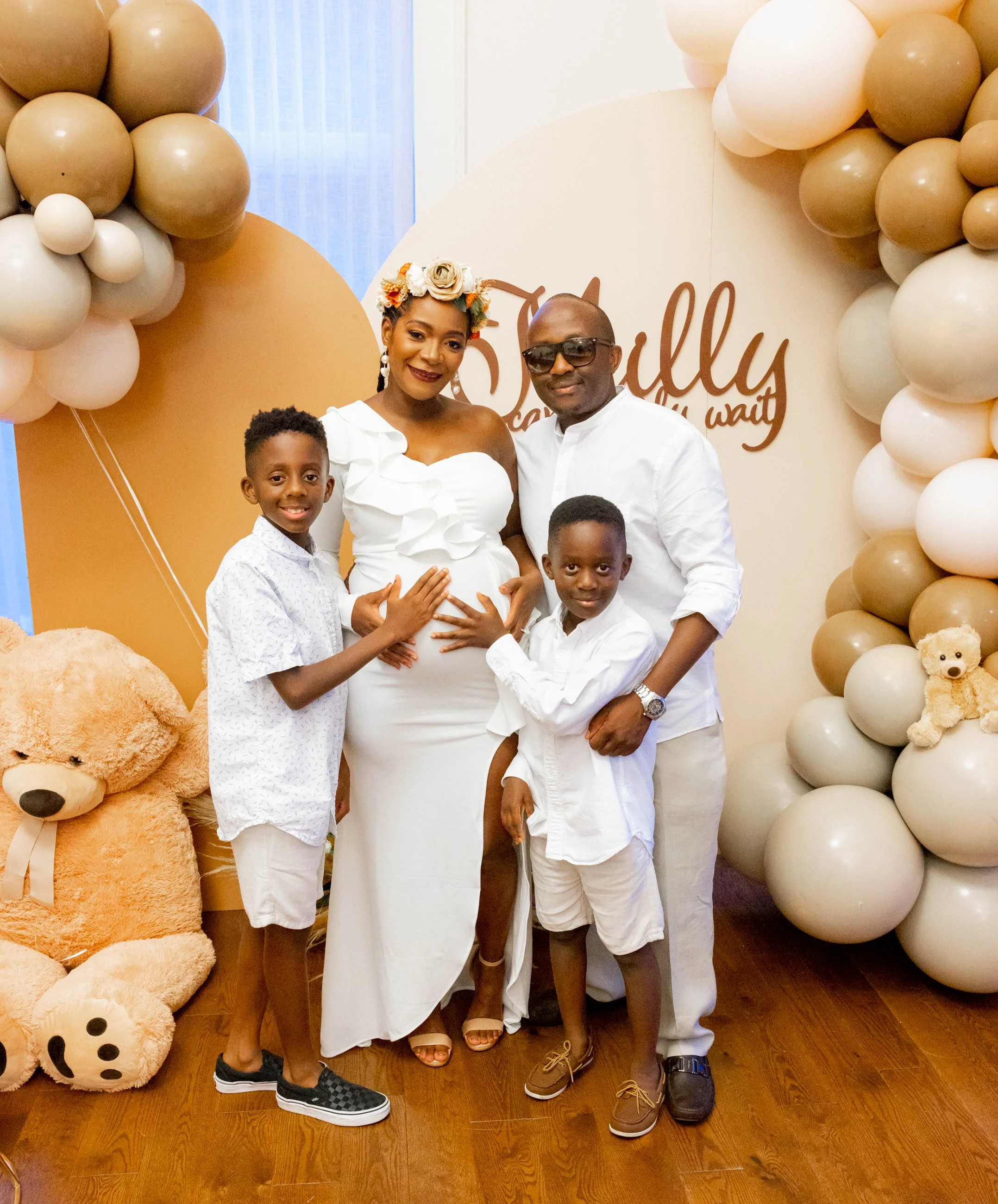 A family portrait with a pregnant woman, two young boys, and a man, standing in front of a balloon decoration for a celebration.