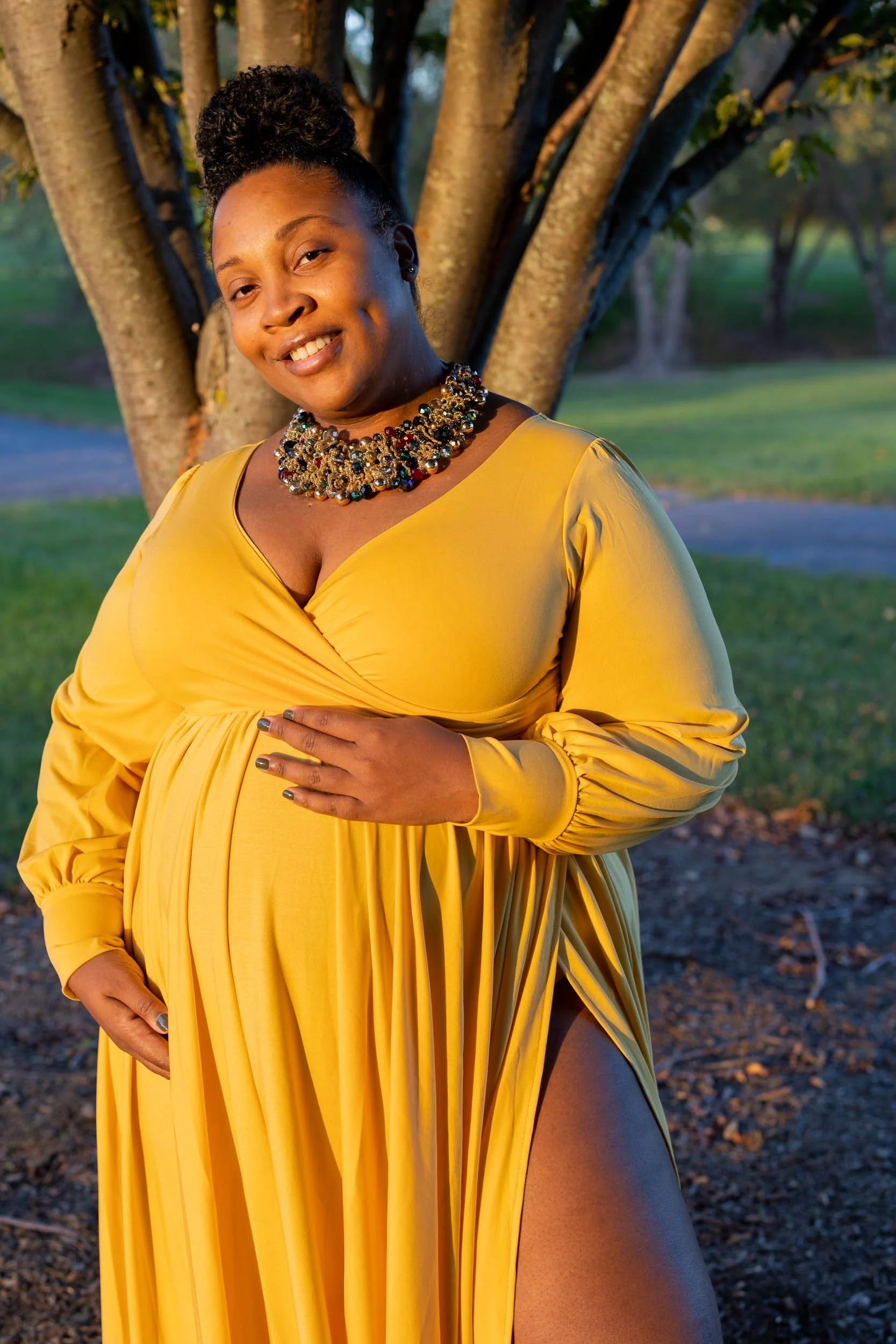 A woman in a long, vibrant yellow dress with long sleeves, standing outdoors in front of a tree. She has her right hand resting on her stomach and her left hand down by her side. She is smiling and wearing an elaborate, colorful beaded statement neck