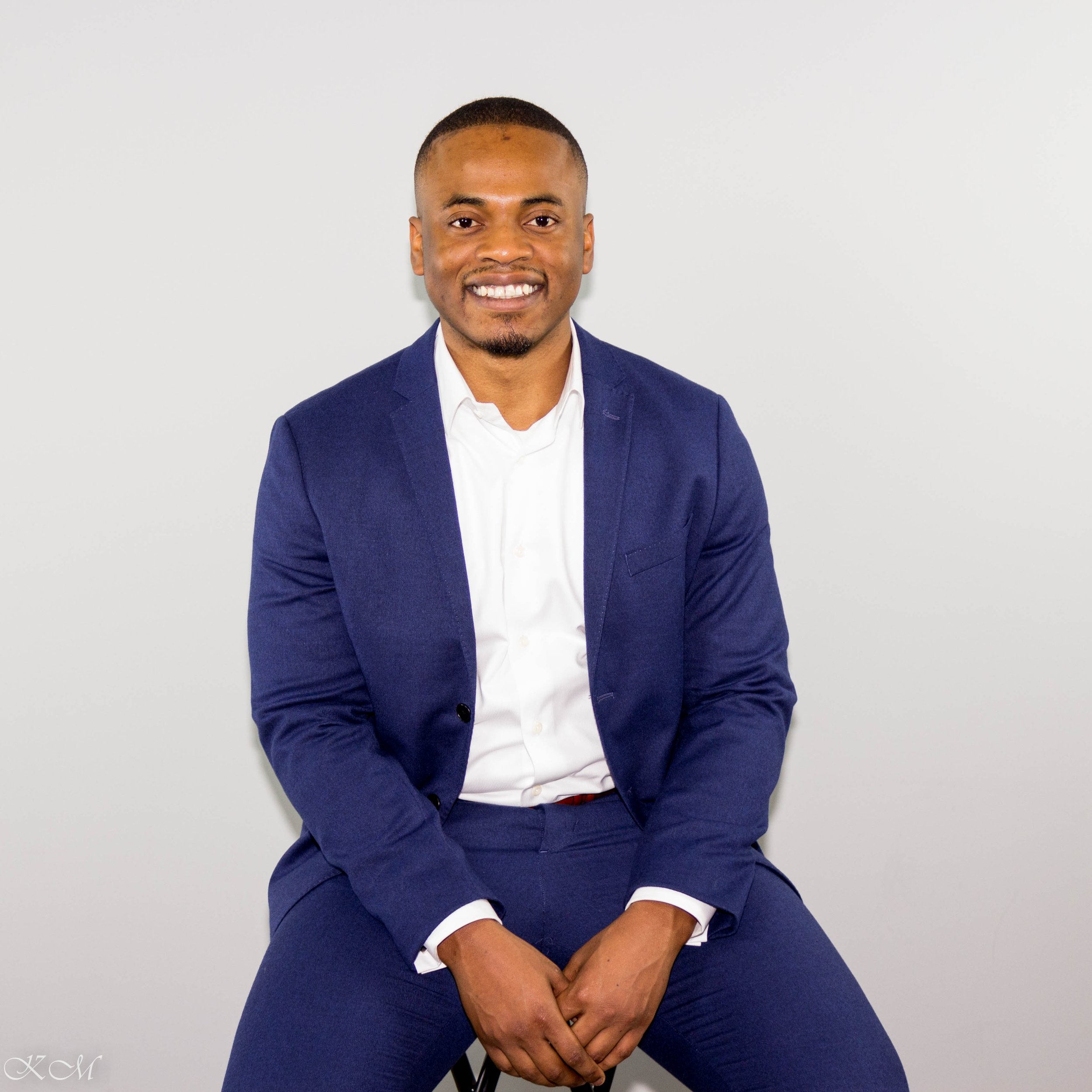 A smiling man in a navy blue suit and white shirt sitting against a plain white background.