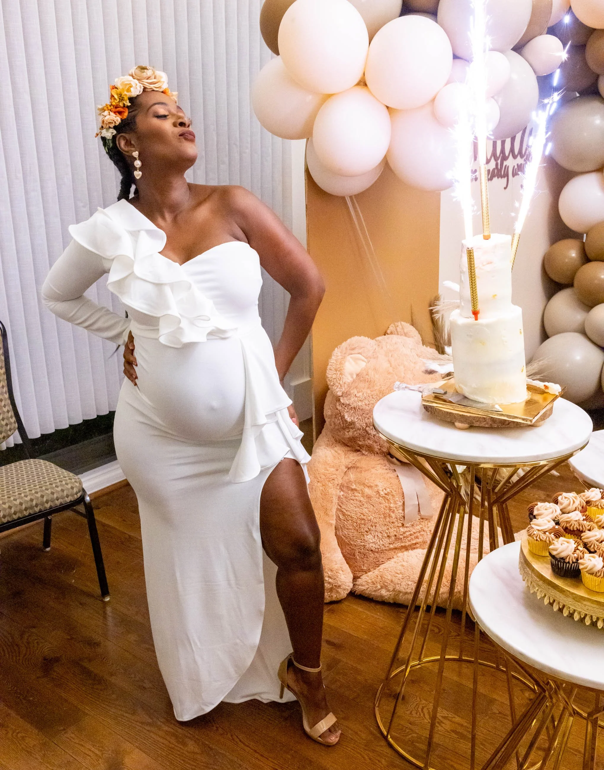 A woman in a white dress with a high slit and ruffle details stands near a birthday cake with sparklers, celebrating at a decorated party with balloons and a large pink teddy bear in the background.