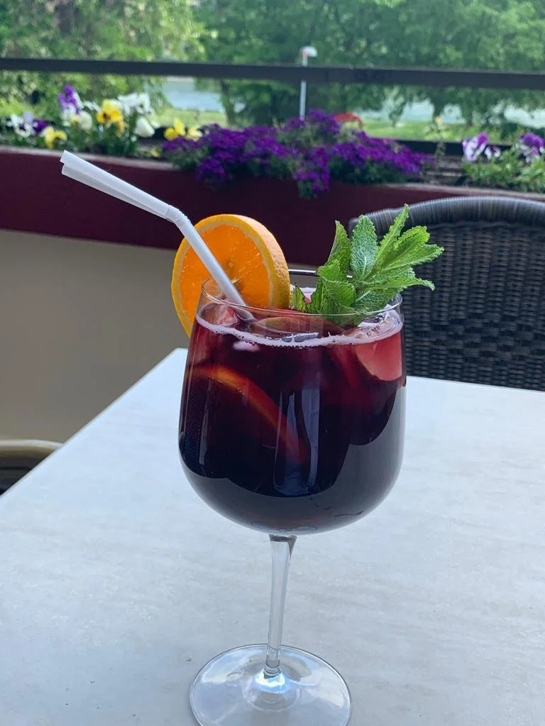 Glas mit Sangria, Apfel und Orange, auf einem weißen Tisch, im Hintergrund Blumen und grüne Bäume.