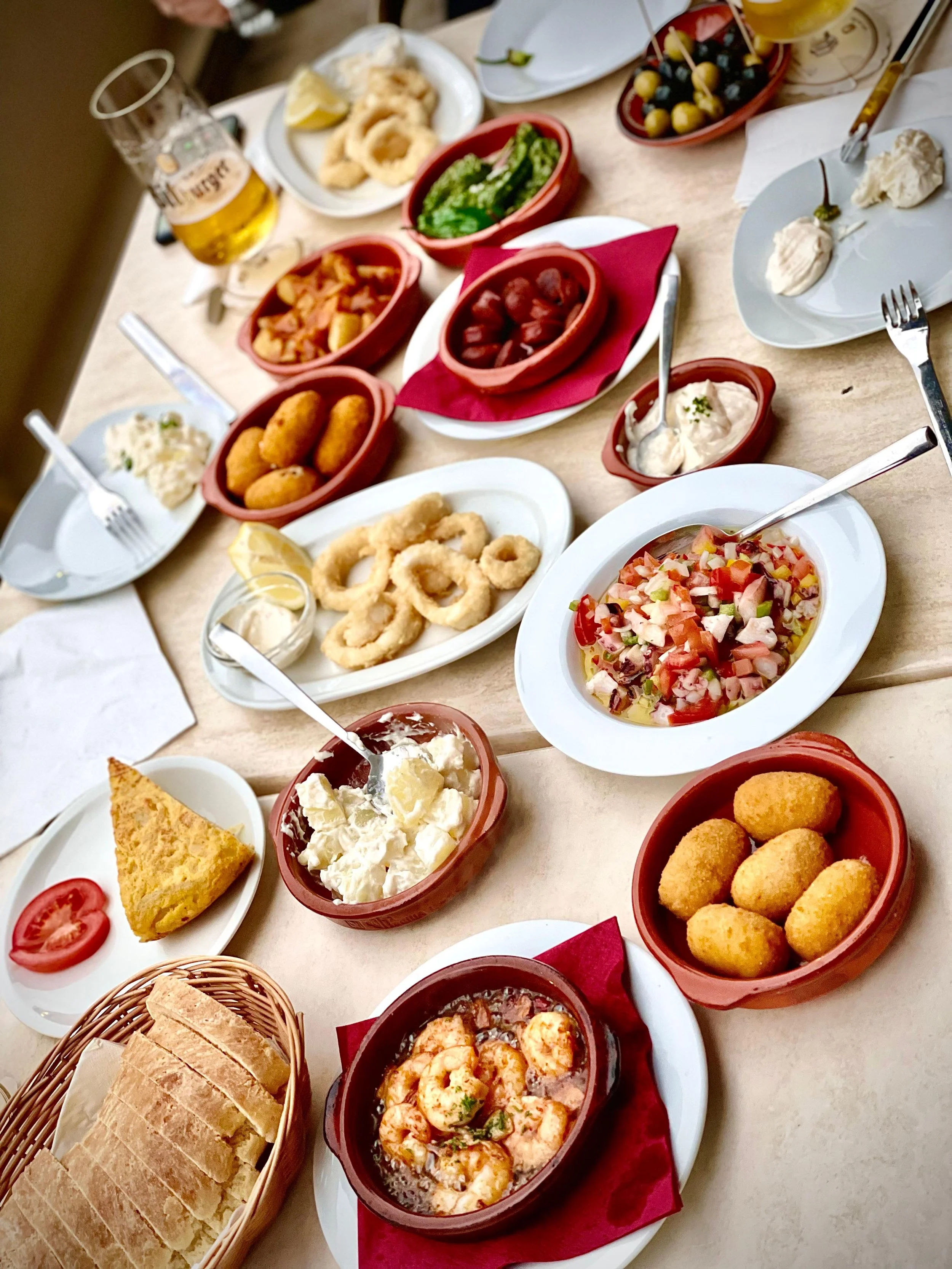Ein Tisch mit spanischen Tapas, darunter Garnelen, Croquetas, Patatas Alioli, Alioli, Patas Bravas, Brot, fritierte Tintenfischringe, Pulpo Vinagreta, Chorizo, Tortilla und Pimientos. Es gibt auch Gläser mit Bier.