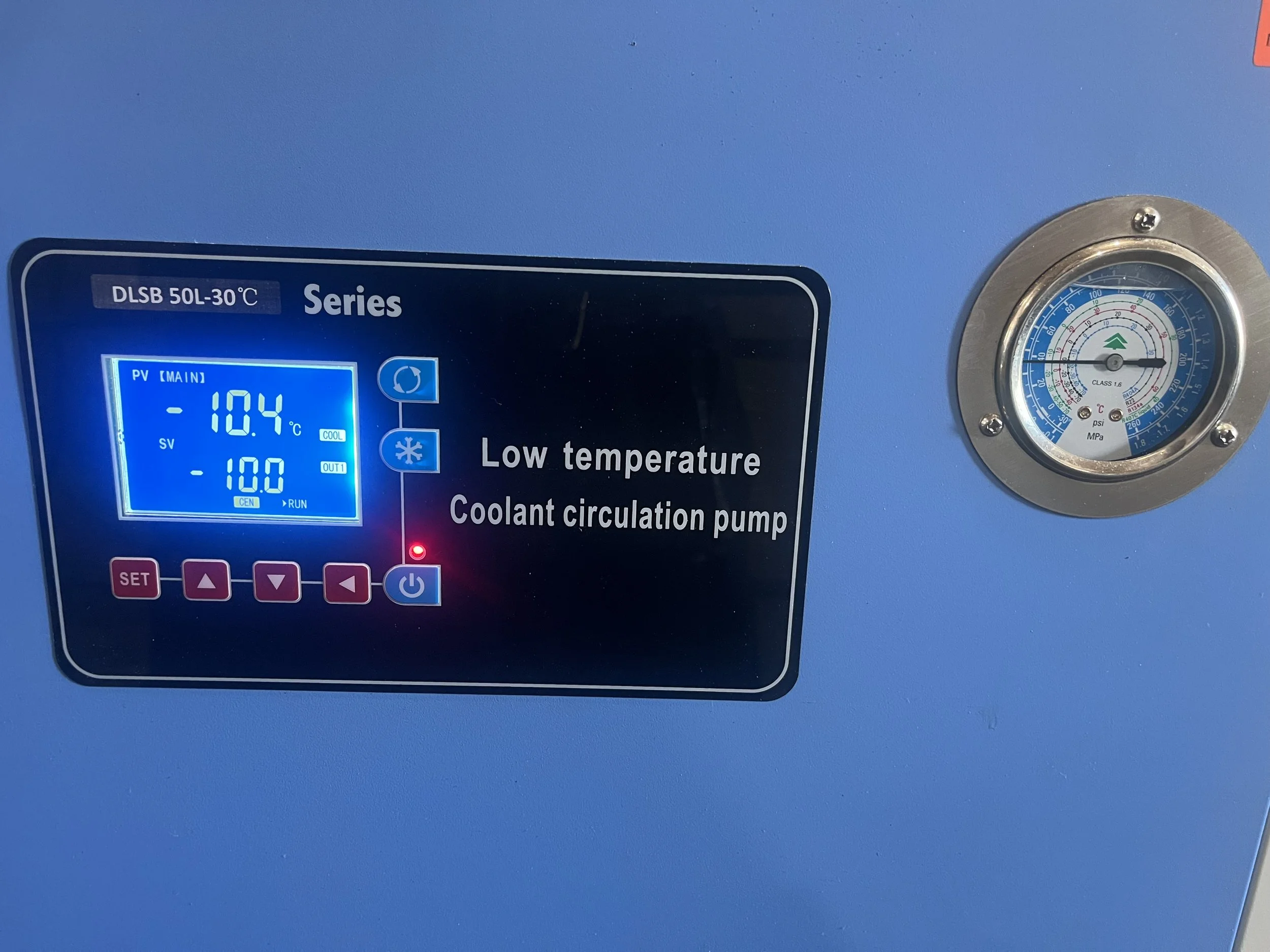 Close-up of a blue industrial cooling machine displaying a temperature of -10.4°C, with a pressure gauge mounted on a light blue wall.