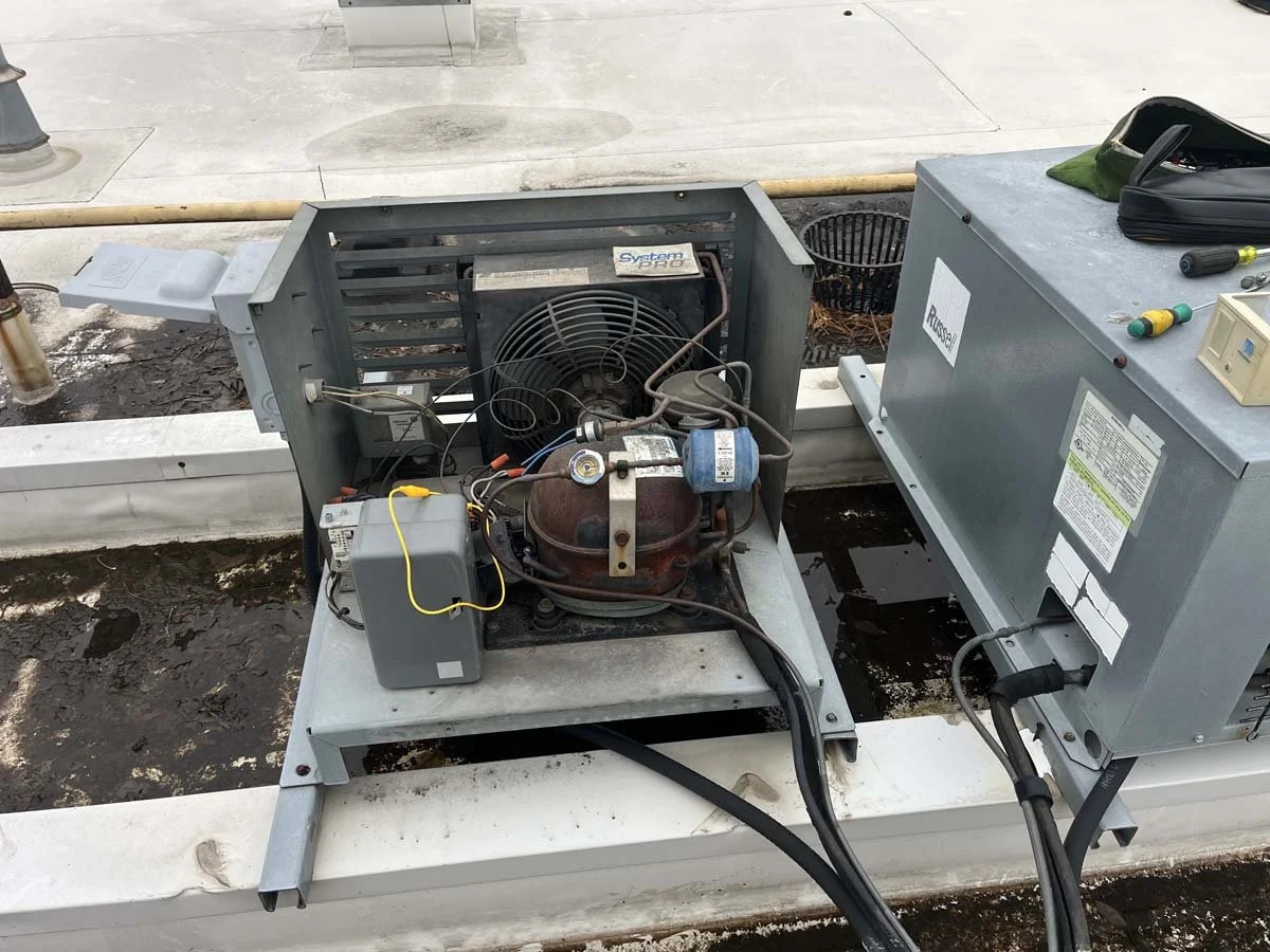 Open HVAC unit with compressor and electrical components, placed on a rooftop with tools and equipment nearby.