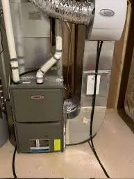 A utility room with a gas furnace and an electric water heater, with pipes and electrical wiring visible on a wooden wall.