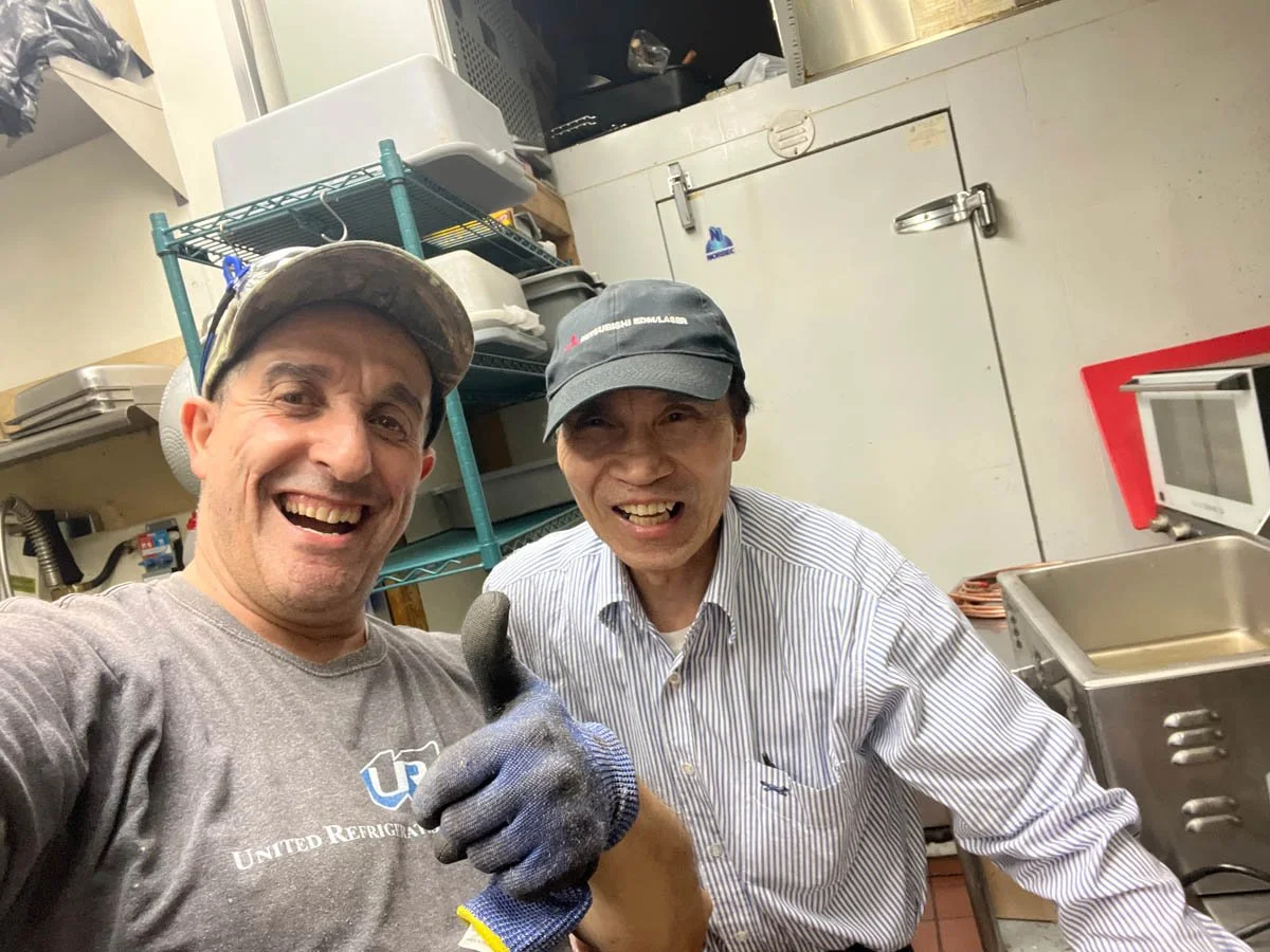 Two men smiling in a kitchen, one showing a thumbs-up and the other slightly grimacing. Both are wearing work gloves and caps.