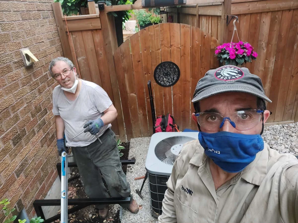 Two men working in a backyard enclosed by a wooden fence, one wearing a cap, glasses, and a face mask, taking a selfie, and the other with gloves, smiling, holding gardening tools, with a flower hanging on the fence and a small air conditioning unit 