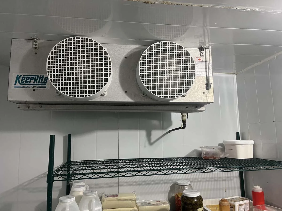 An industrial refrigeration unit mounted on a wall with KeeRite branding, metal wire shelving below with food containers and bottles, in a kitchen or storage room.