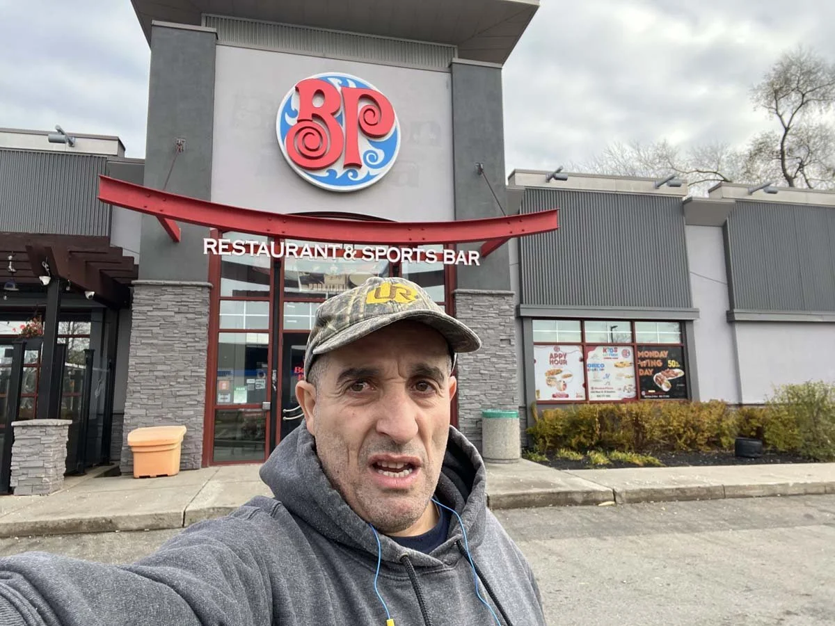 Man taking a selfie outside a restaurant and sports bar. The building has a large colorful circular sign with a red and blue design and the word 'BPU' or similar. The man wears a gray hoodie and a camouflage cap, appears tired or confused.