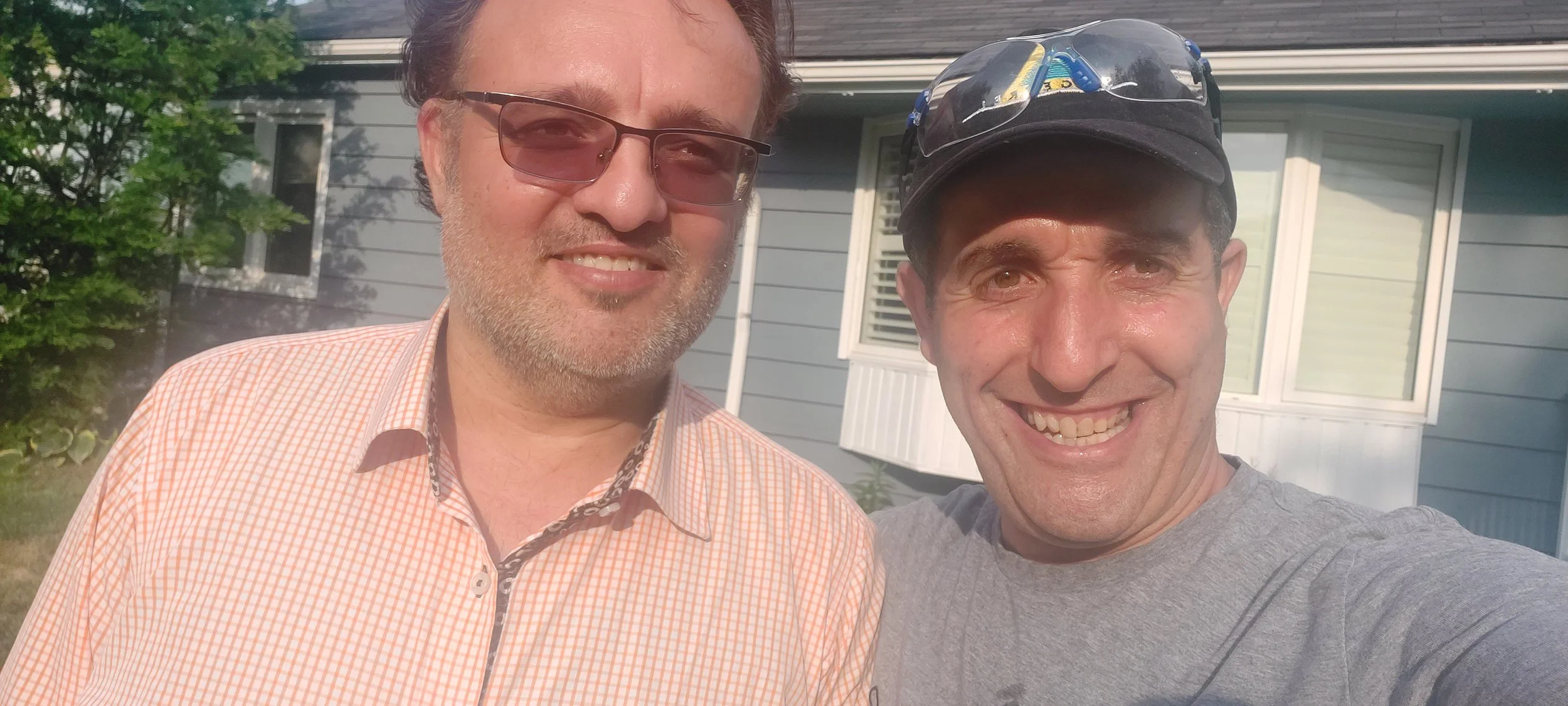 Two men smiling and taking a selfie outside in front of a house, one wearing glasses and a checked shirt, the other wearing a cap and gray t-shirt.