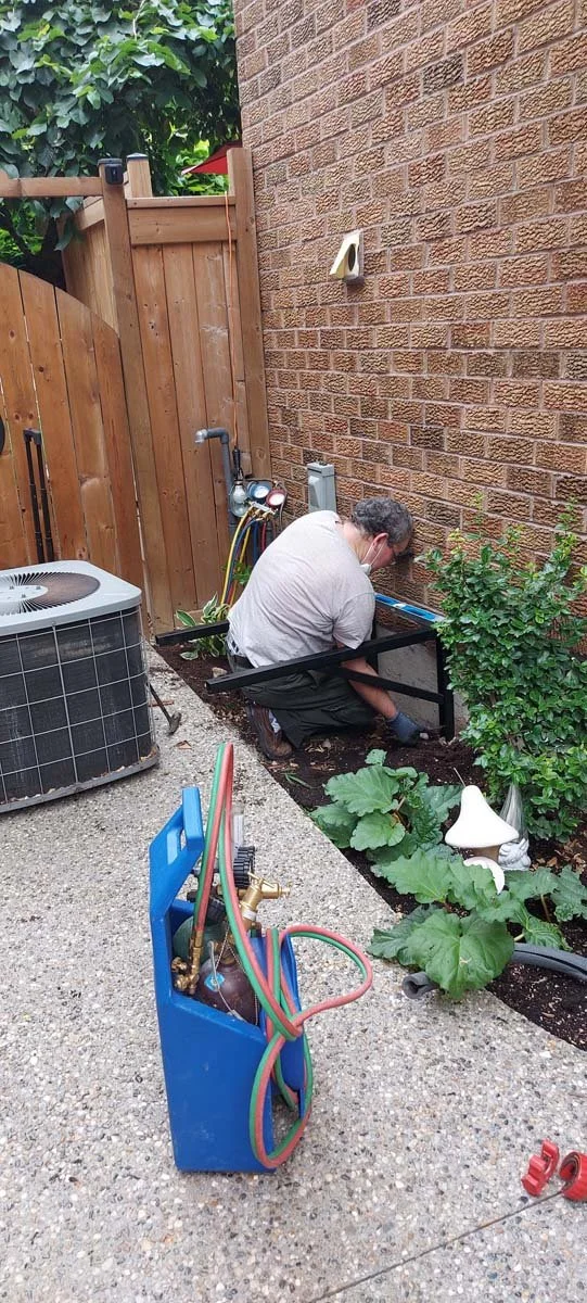 A man wearing a face mask working on gardening or plumbing near a brick wall and wooden fence, with HVAC equipment and tools around.