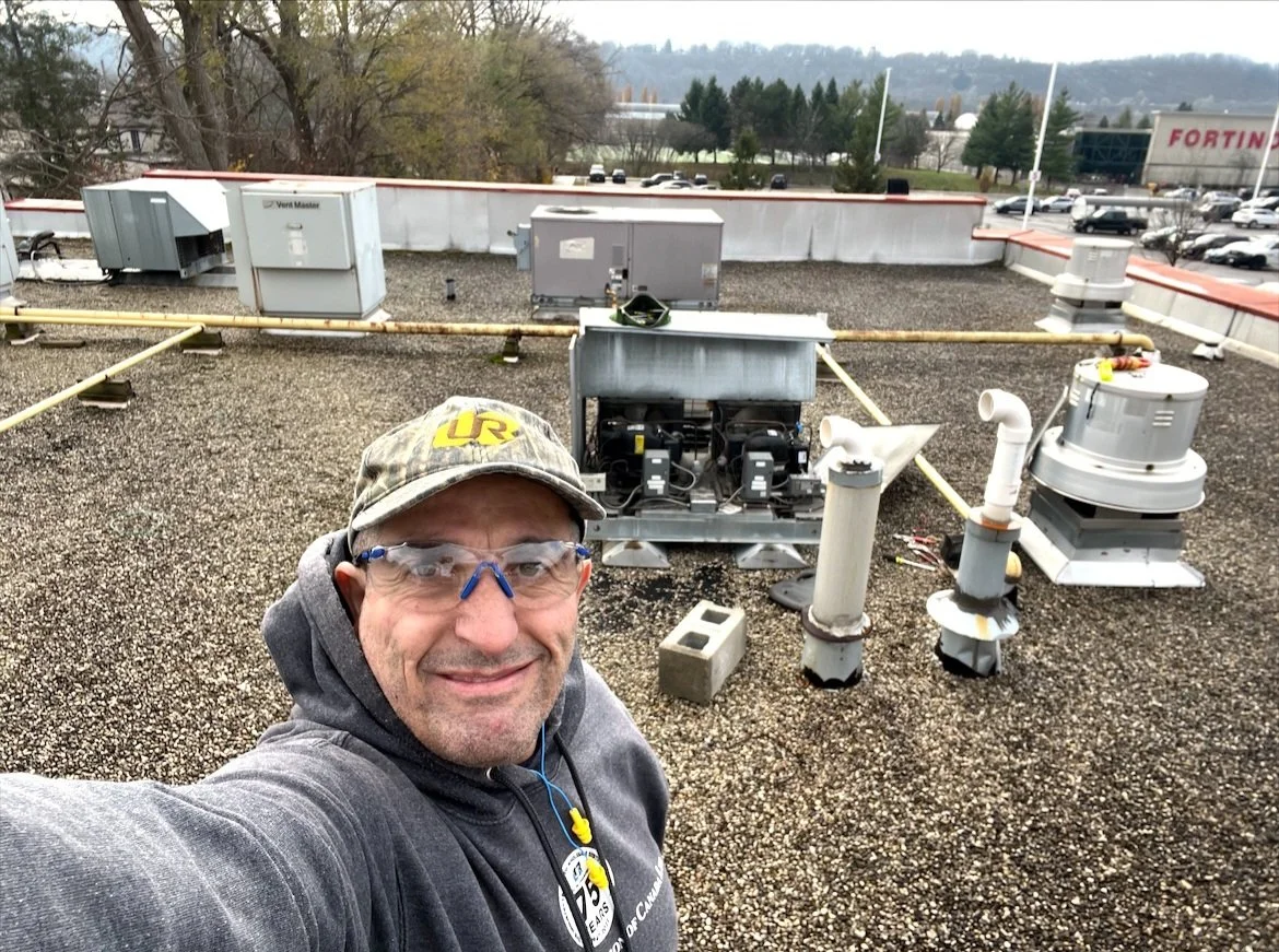 Man taking a selfie on a building rooftop with HVAC and ventilation equipment, parking lot, and shopping center in the background.