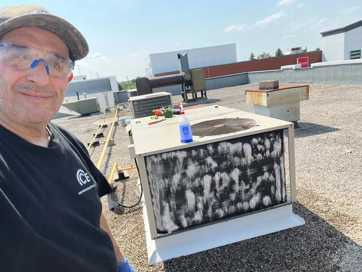 A man wearing safety glasses and a black shirt standing on a rooftop with HVAC units and equipment, including a molecular, spray bottle, and tools, under a partly cloudy sky.