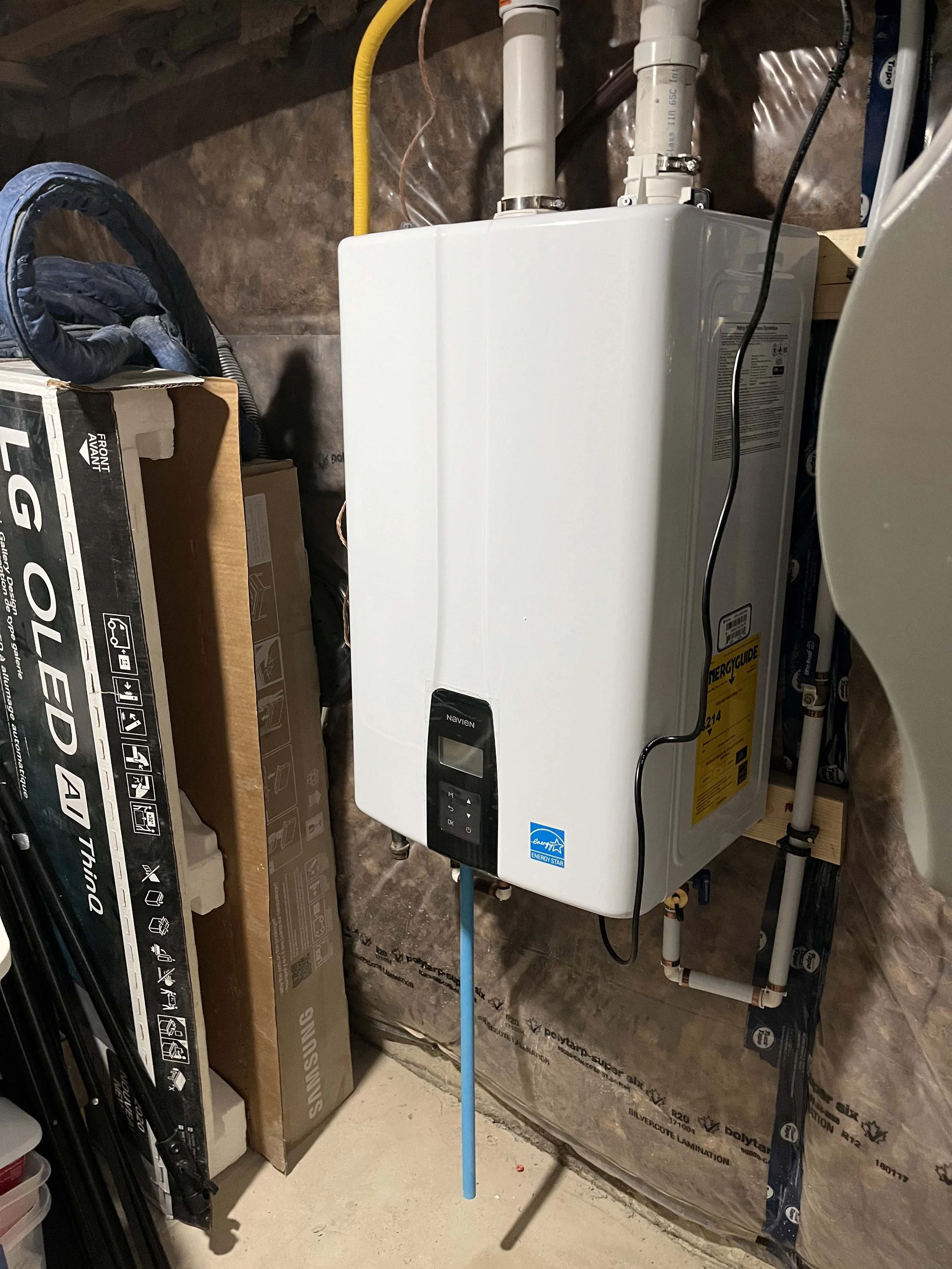 A water heater in a basement or utility room with PVC pipes connected to it. There is a black power cord plugged into the water heater. The room has unfinished walls with insulation and plumbing pipes visible. A blue pole is leaning against the water