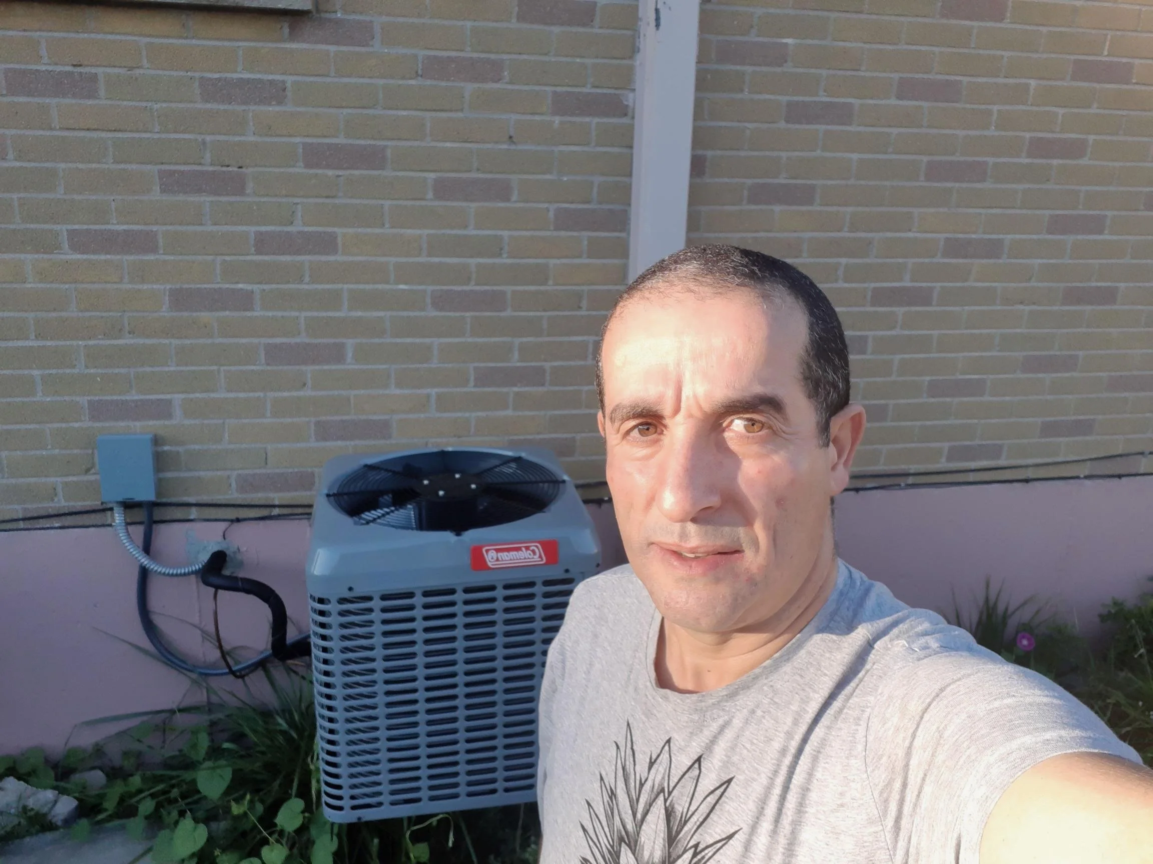 A man with short dark hair and light skin taking a selfie outdoors in front of a brick wall, with an air conditioning unit and some plants nearby.
