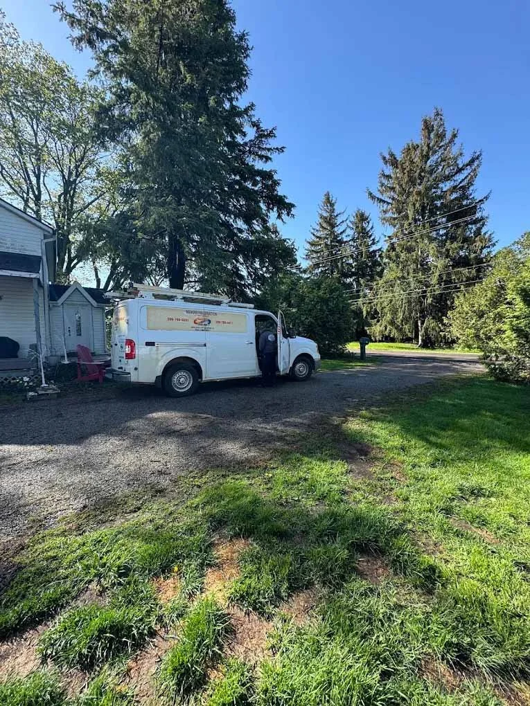 A white service van parked in a driveway near a house on a sunny day, with trees and a clear blue sky in the background.