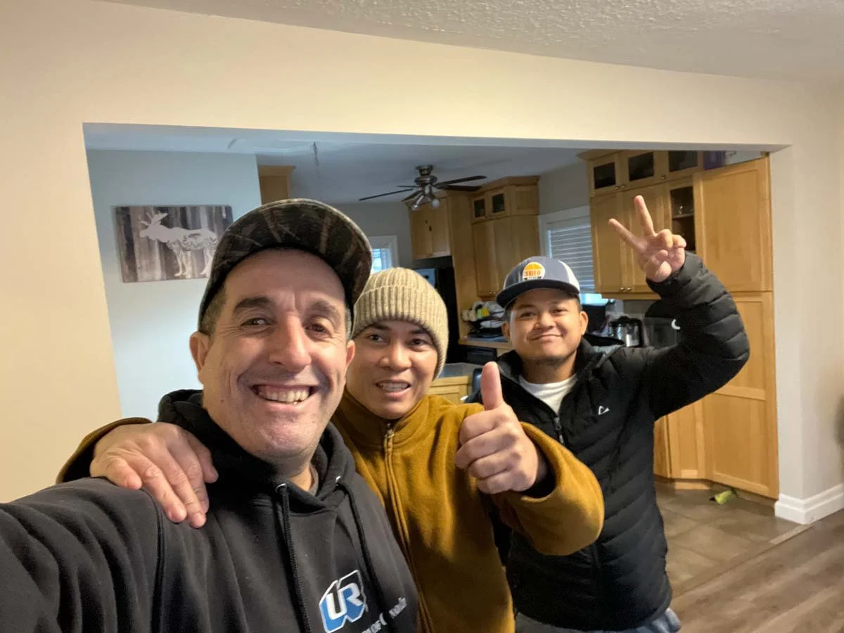 Three smiling men taking a selfie in a kitchen. One man is wearing a black hoodie with a logo, another is in a mustard-colored jacket and beanie, and the third is in a black jacket and cap. The man on the right is making a peace sign with his hand.