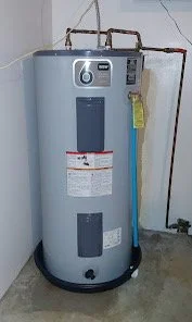 A large industrial water heater or boiler in a utility room, with pipes connected at the top and a yellow tag hanging from one pipe.