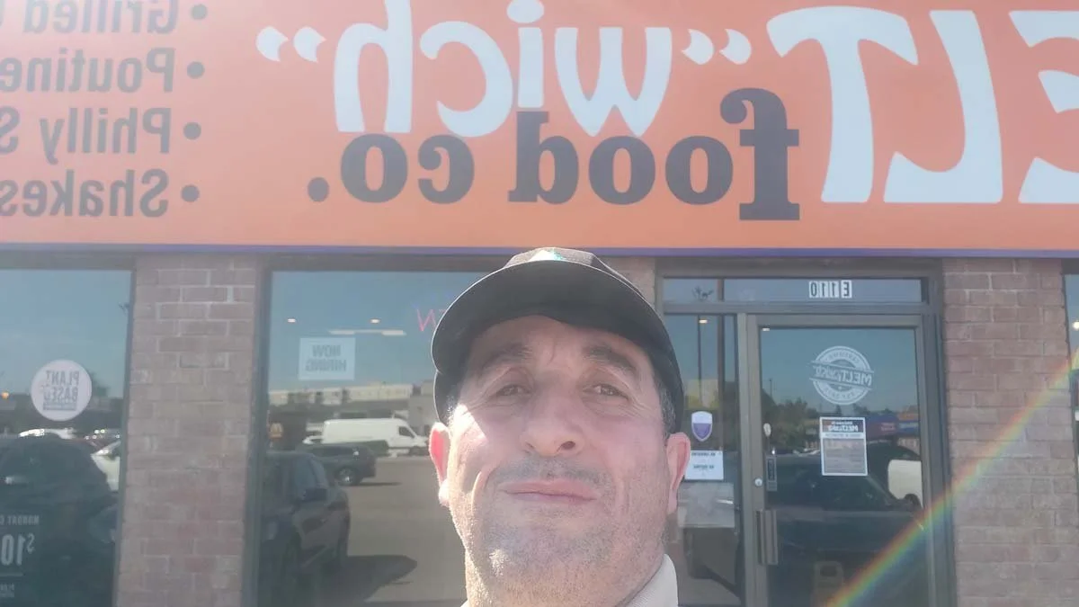 A man taking a selfie outside a food restaurant with large orange and white signage on the building.