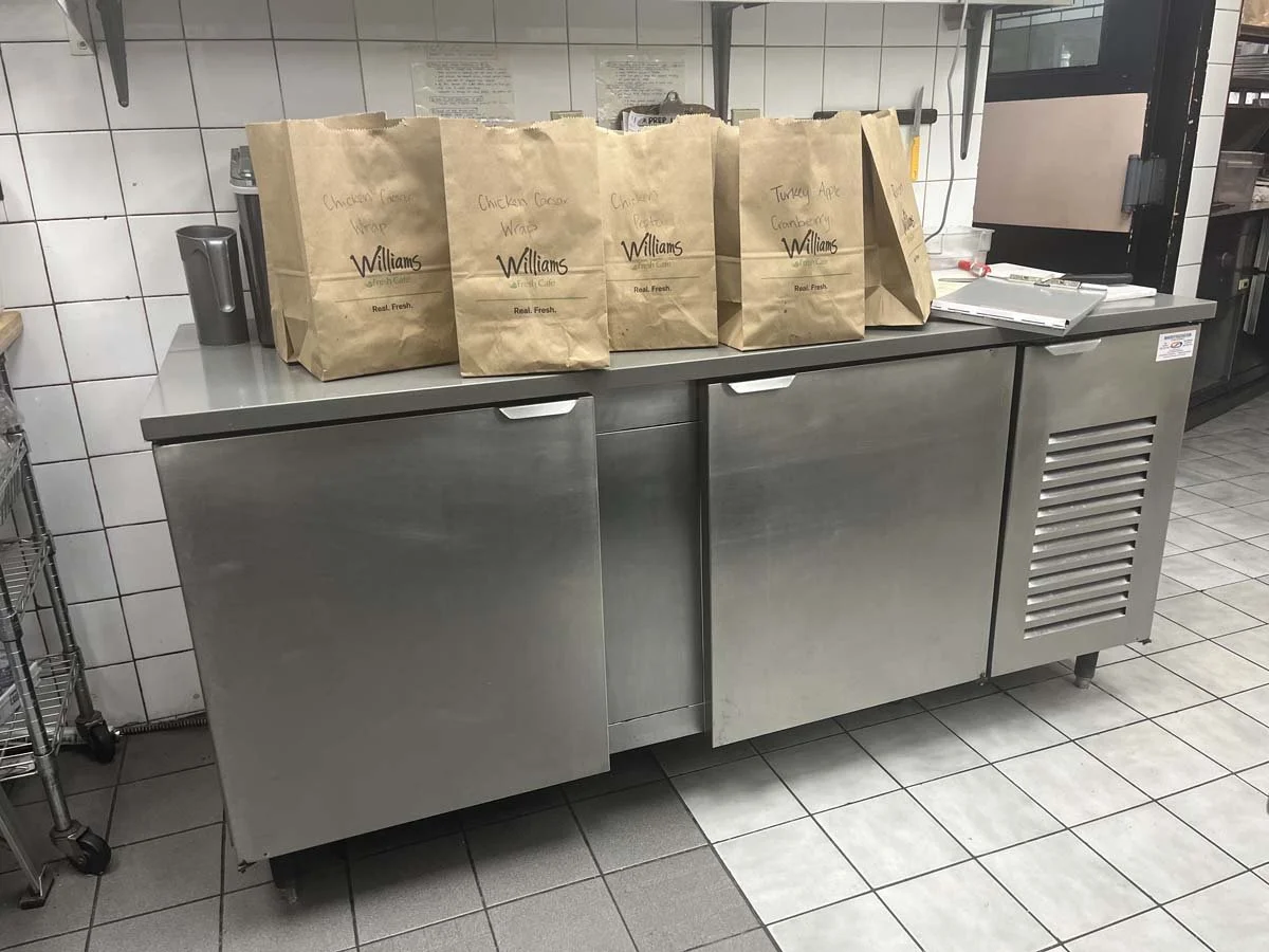 Stainless steel commercial refrigerator with paper bags on top labeled with different sandwich types, in a kitchen with tiled walls and floor.