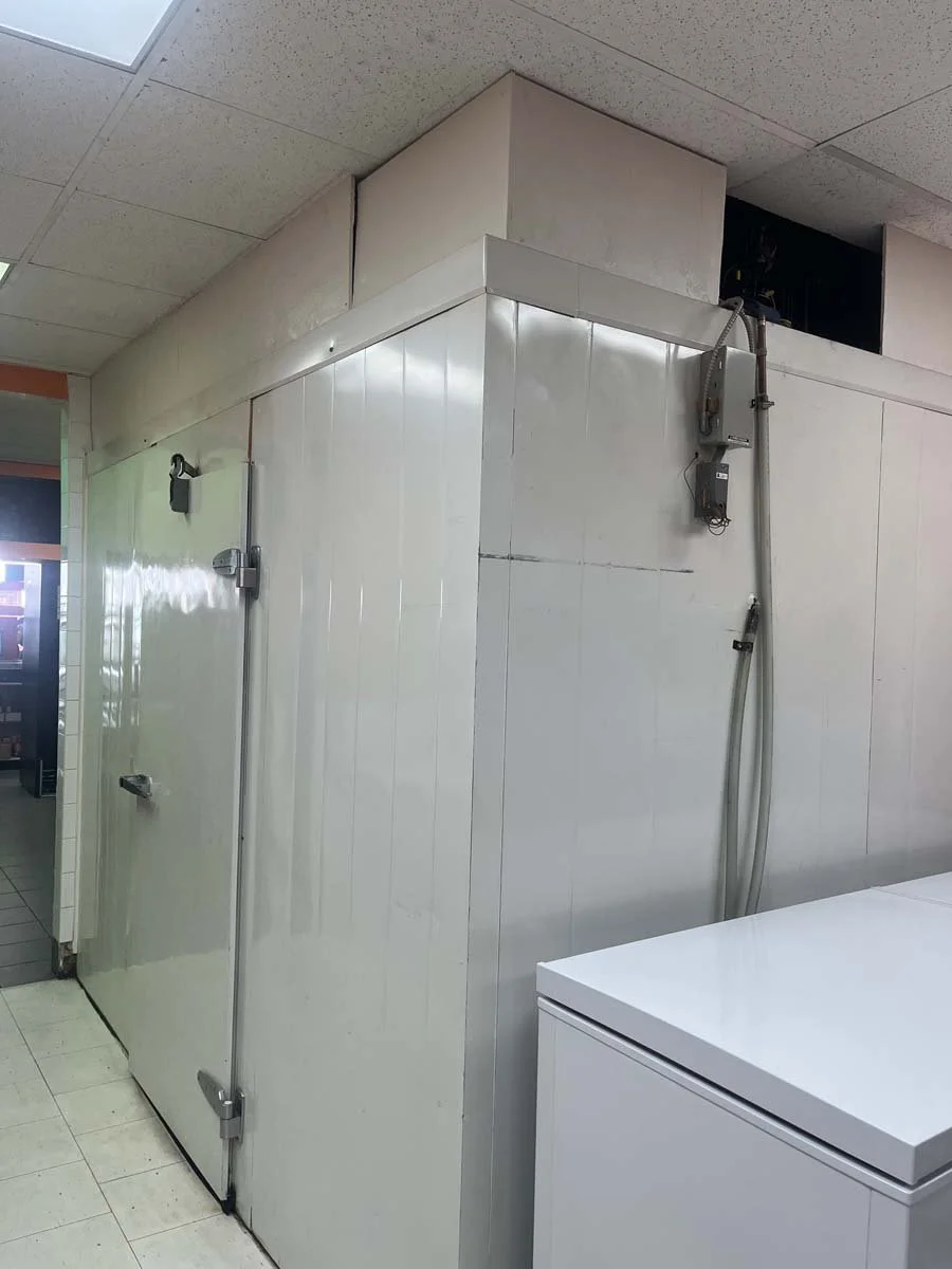 An industrial cold storage or walk-in freezer with a large metal door, hinges, and a latch. There are electrical wires and equipment mounted on the wall beside it. Part of a white commercial appliance is visible in the foreground.