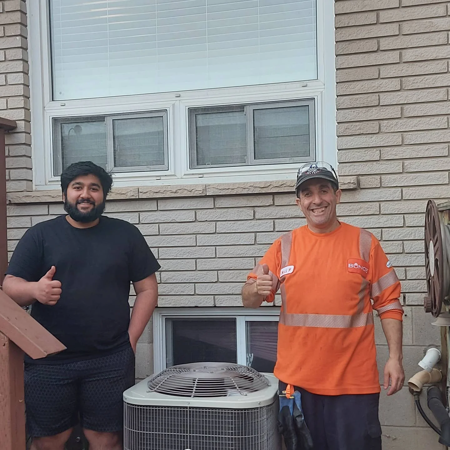 Two men standing outside in front of a brick house wall, giving thumbs up. One wearing a black t-shirt and shorts, the other in an orange work shirt with reflective stripes, a cap, and a tool pouch.