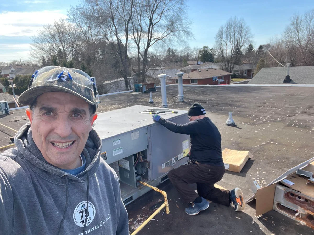 Two men working on an HVAC unit on a rooftop in a residential neighborhood, with leafless trees and houses in the background.