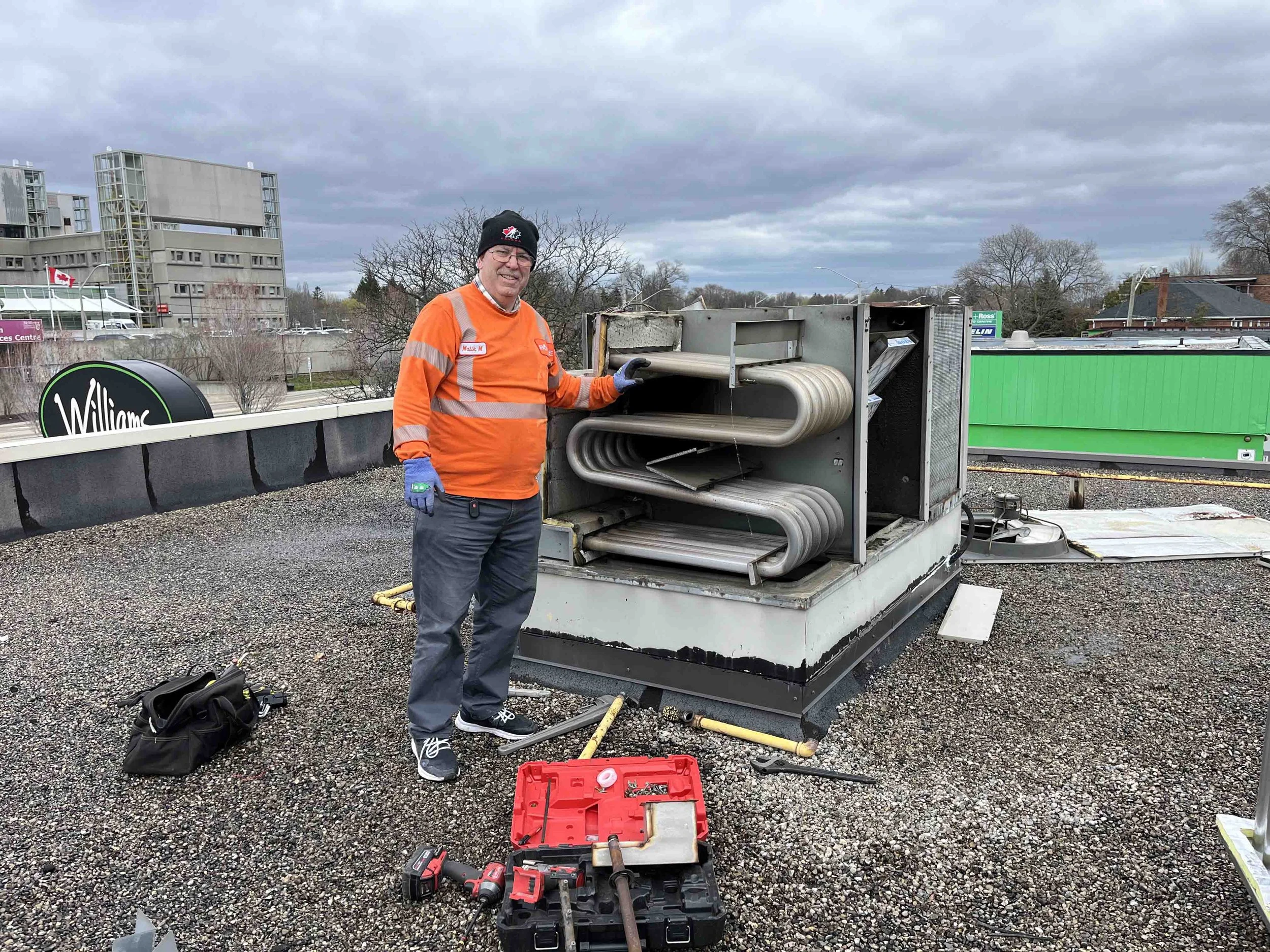 A man wearing glasses, a black beanie, and an orange safety jacket stands on a rooftop with tools, including a red toolbox, power drills, and other equipment, in front of a large HVAC unit or industrial equipment. In the background, there are buildings, trees, and a cloudy sky.