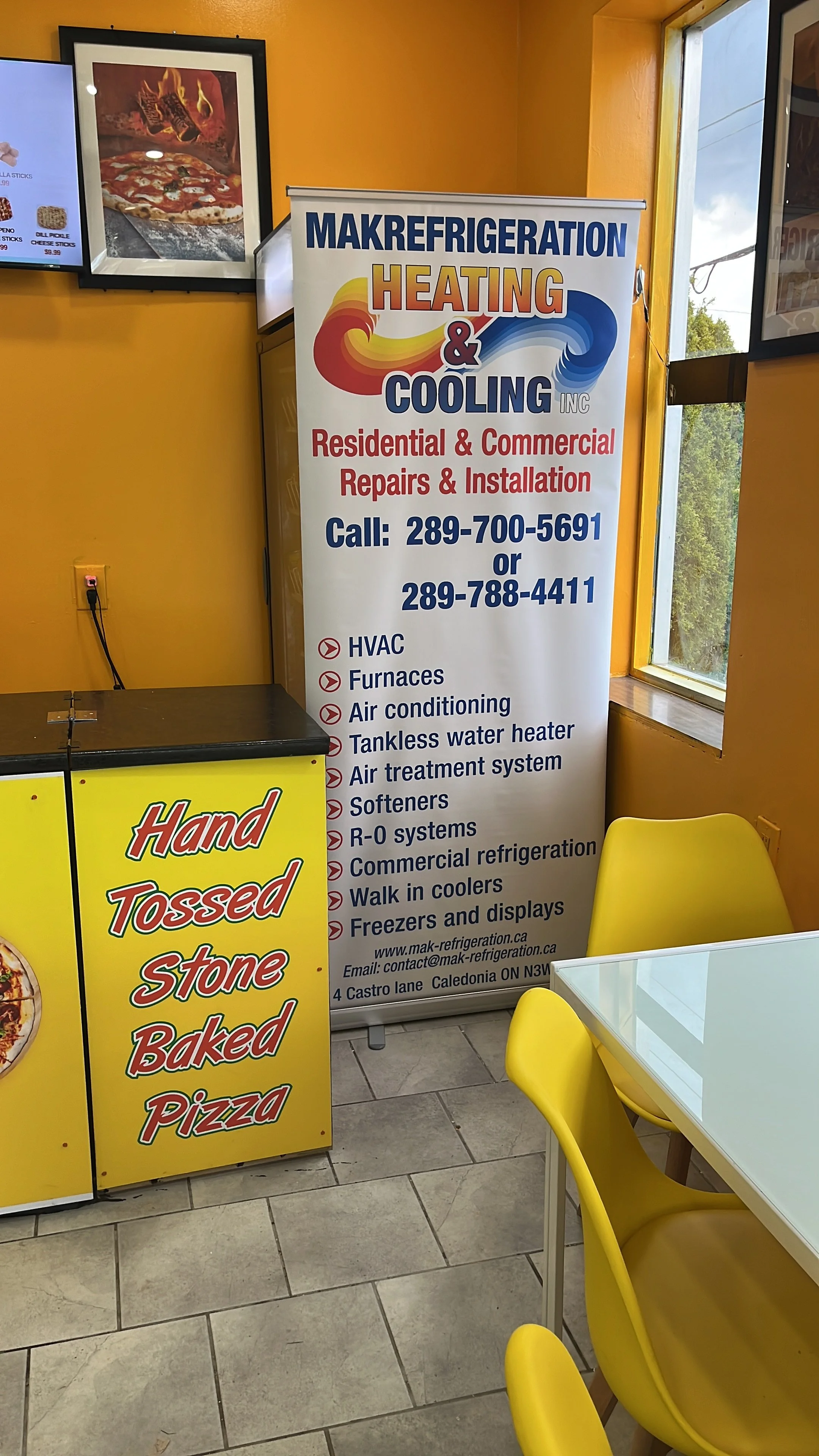 Interior of a restaurant or shop with bright yellow walls, showing a large standing sign for MakRefrigeration Heating & Cooling Inc. listing services such as HVAC, furnaces, air conditioning, and refrigerators, with contact numbers. To the left, a me