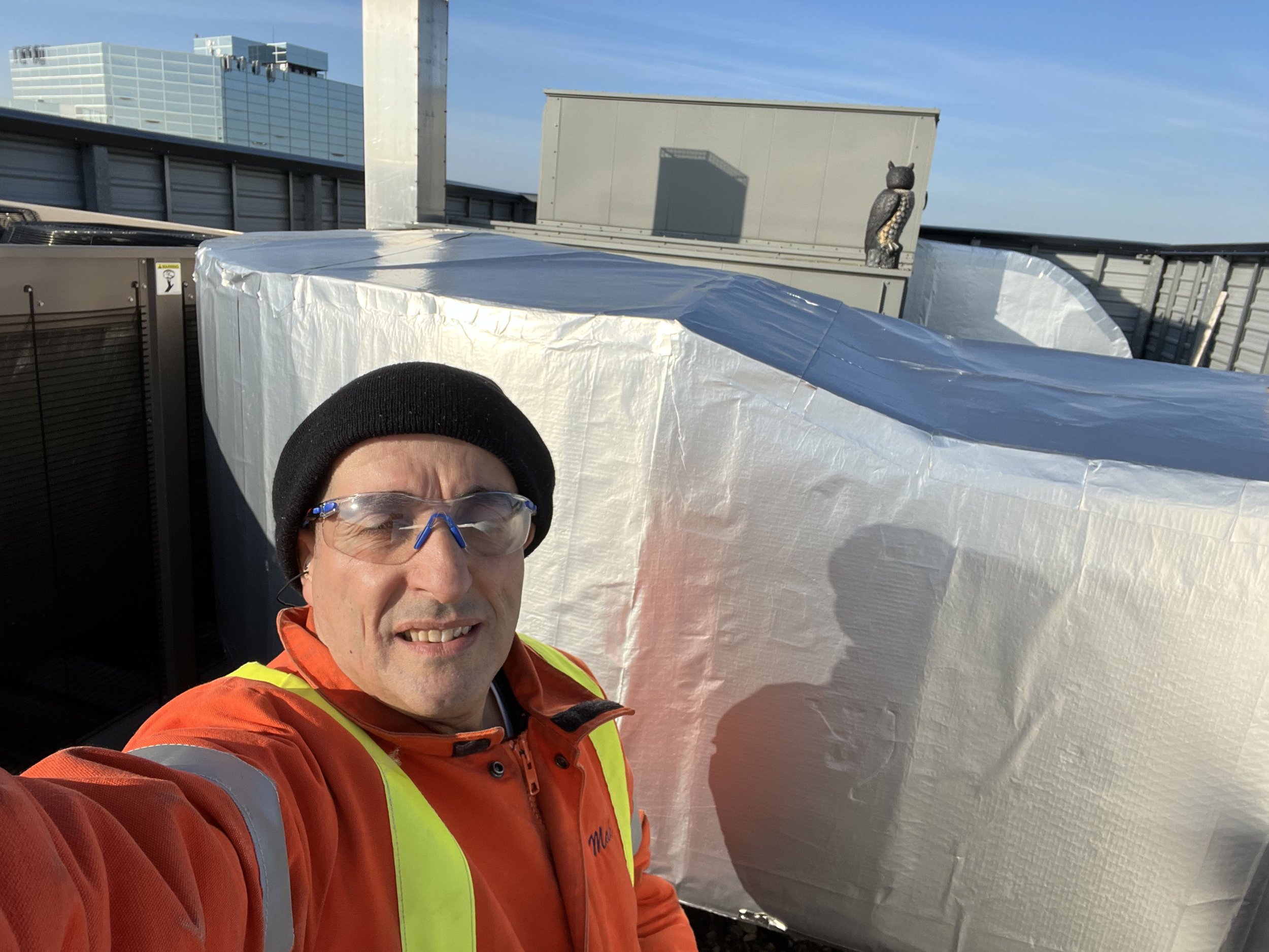A man wearing safety glasses, an orange jacket, and a high-visibility vest in front of a rooftop HVAC unit with a protective covering on it and a rooftop air conditioning unit with a decorative owl statue on top.