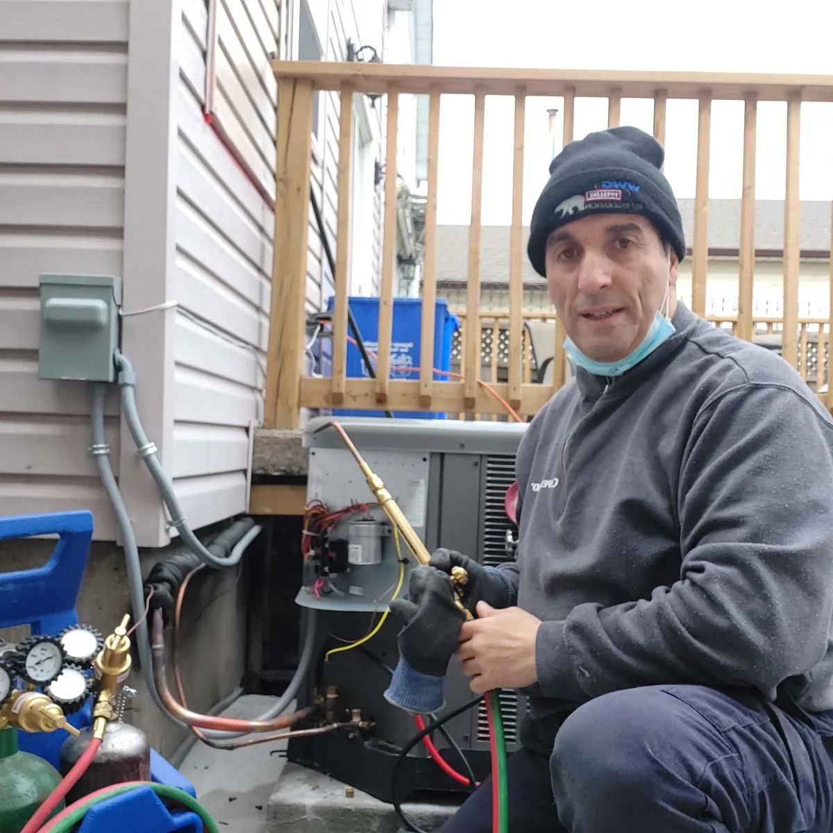 A man in a black beanie and gray hoodie working on an air conditioning unit outside his house. He is holding a refrigerant gauge hose and wearing gloves. There are tools and gauges nearby, with a wooden deck and house siding in the background.