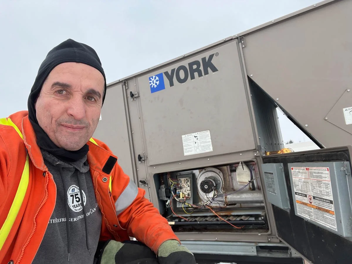 A man wearing an orange jacket, black cap, and gloves taking a selfie next to an industrial York HVAC unit with its service panel open.