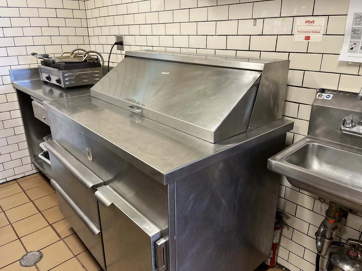 Commercial kitchen with stainless steel appliances, including a flat-top grill, a sink, and a refrigerator, against tiled walls.