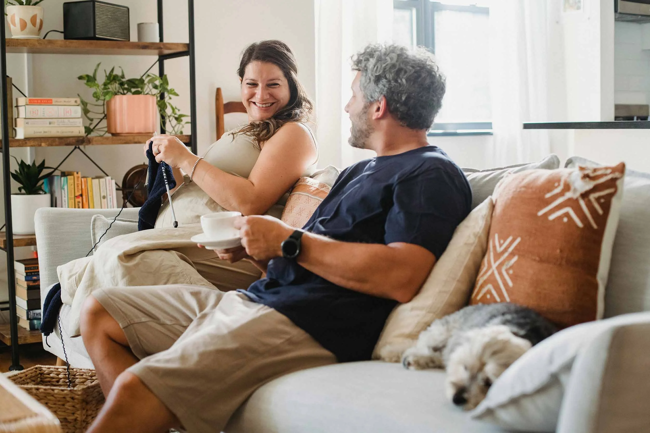 A woman and a man sitting on a grey couch, enjoying each other's company. The woman is knitting while the man has a coffee in his hand.
